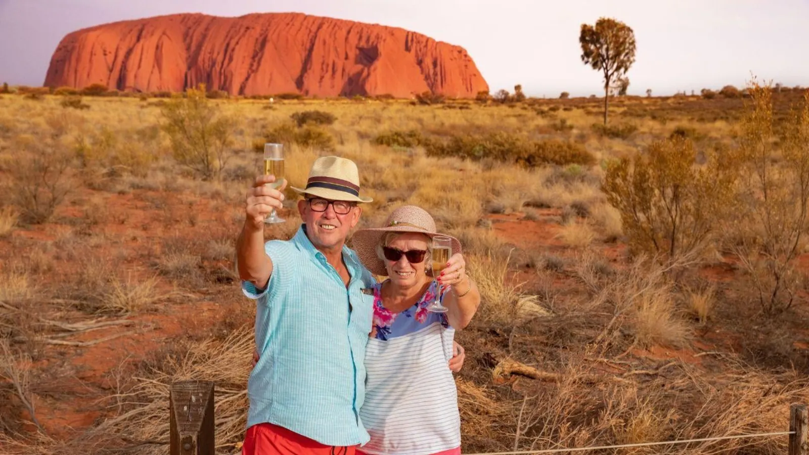Couple in sun hats raising glasses of sparkling wine at the fence with Uluru lit orange