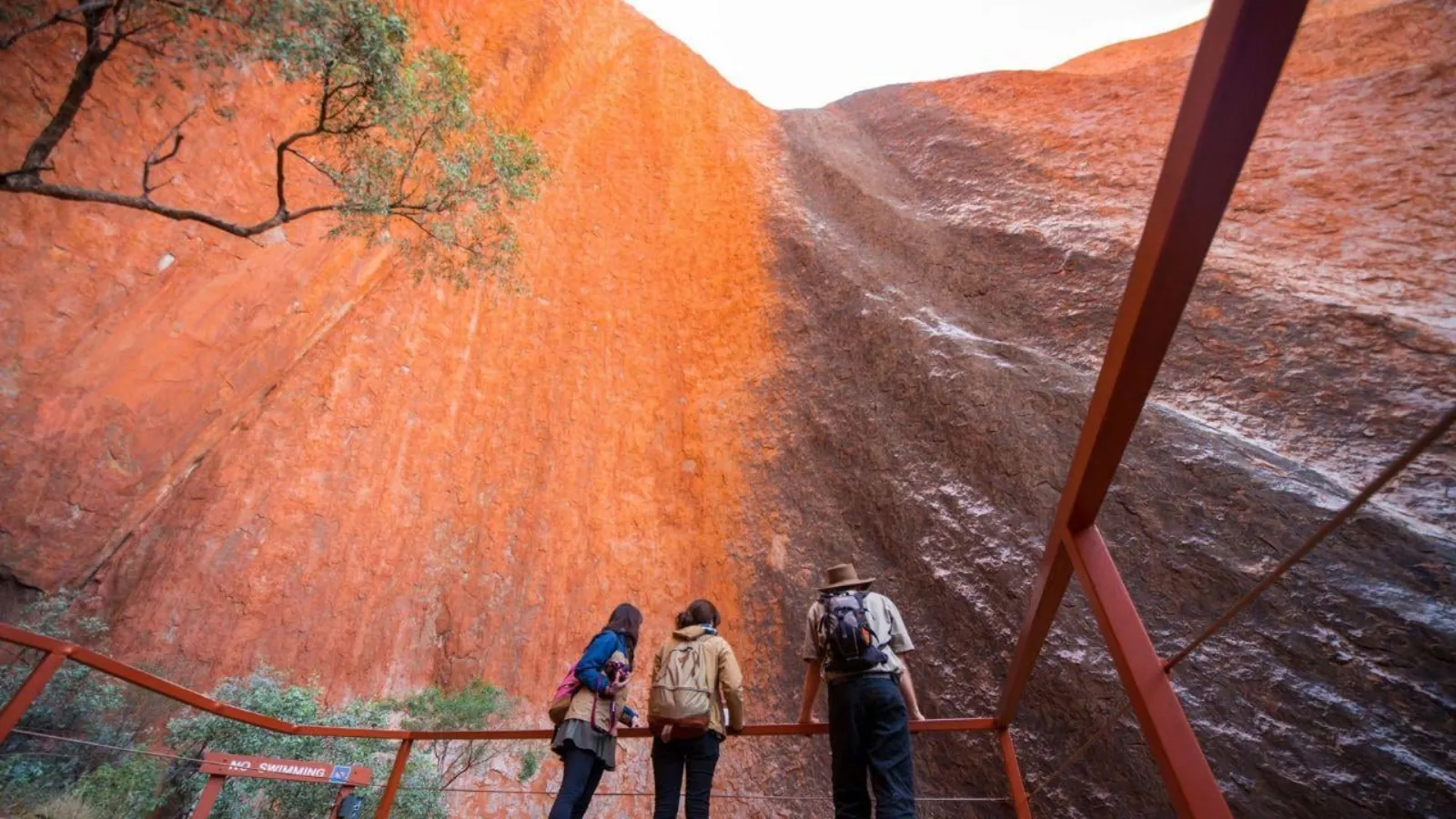 Three walkers at a railing looking up at the streaked rock wall above Mutitjulu Waterhole