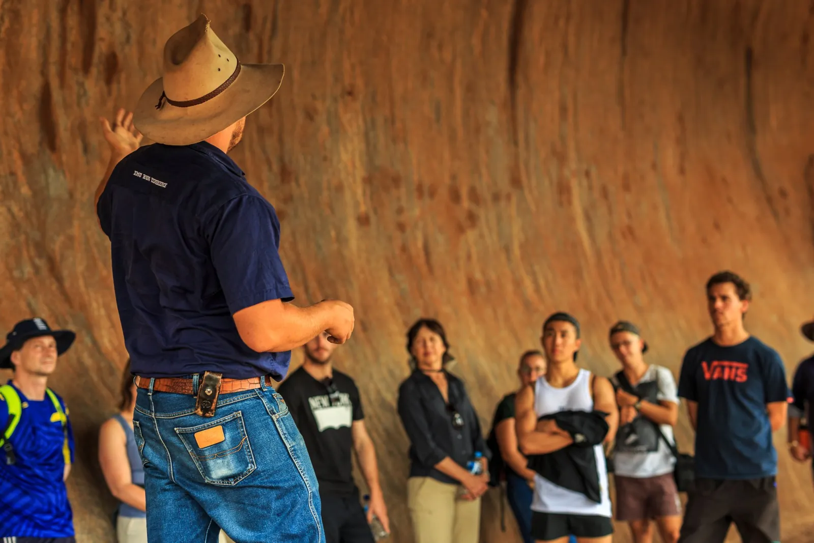Emu Run guide in a bush hat telling a story to a group against Uluru's curved rock wall