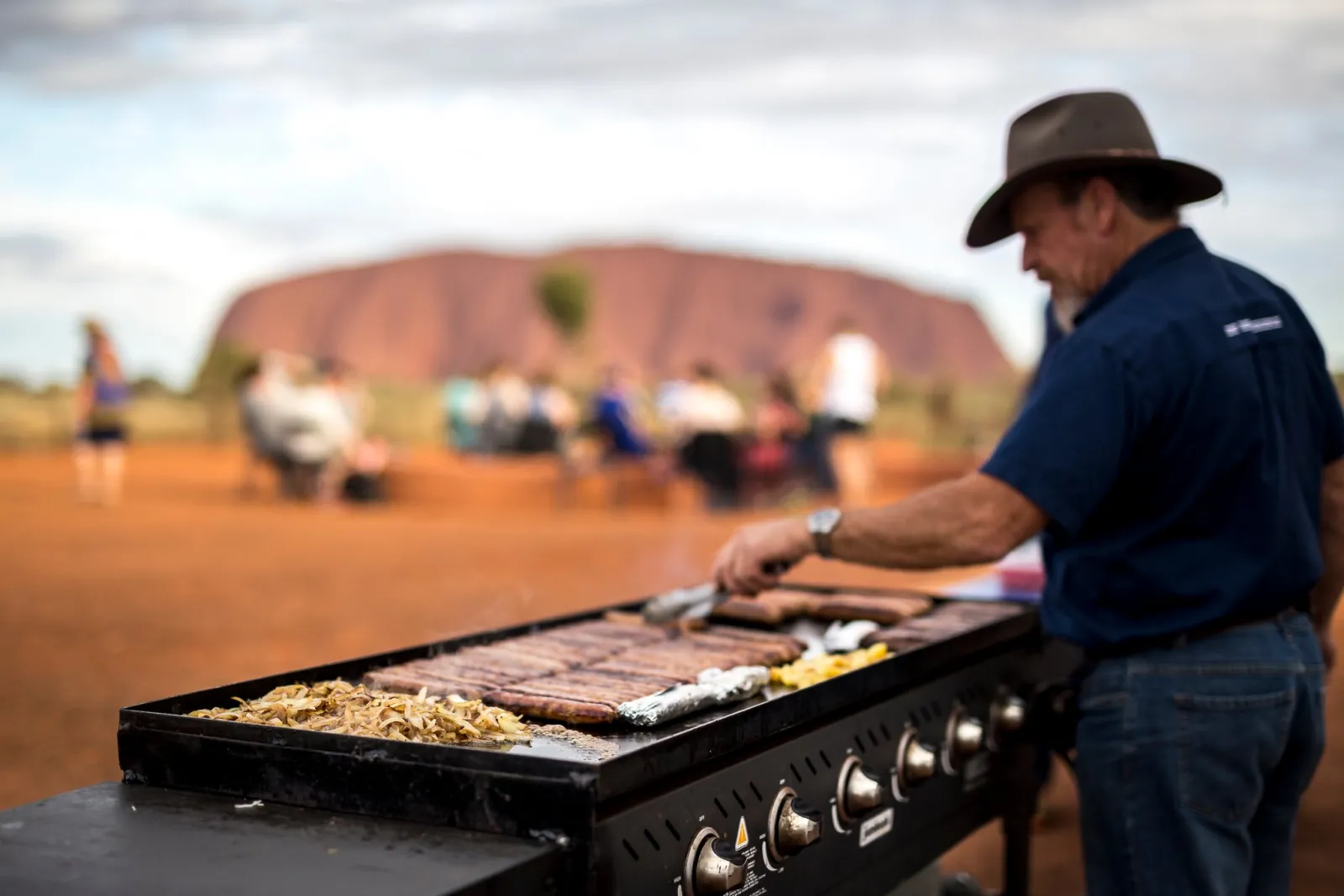 Crew member grilling sausages and onions on a BBQ plate while guests sit near Uluru