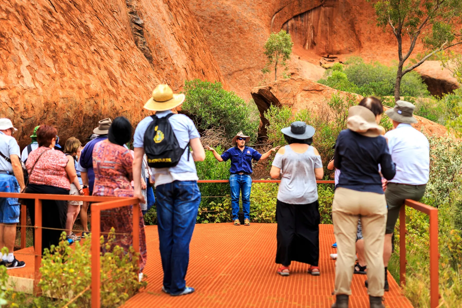 Guide addressing a tour group on an orange viewing platform below Uluru's rock face