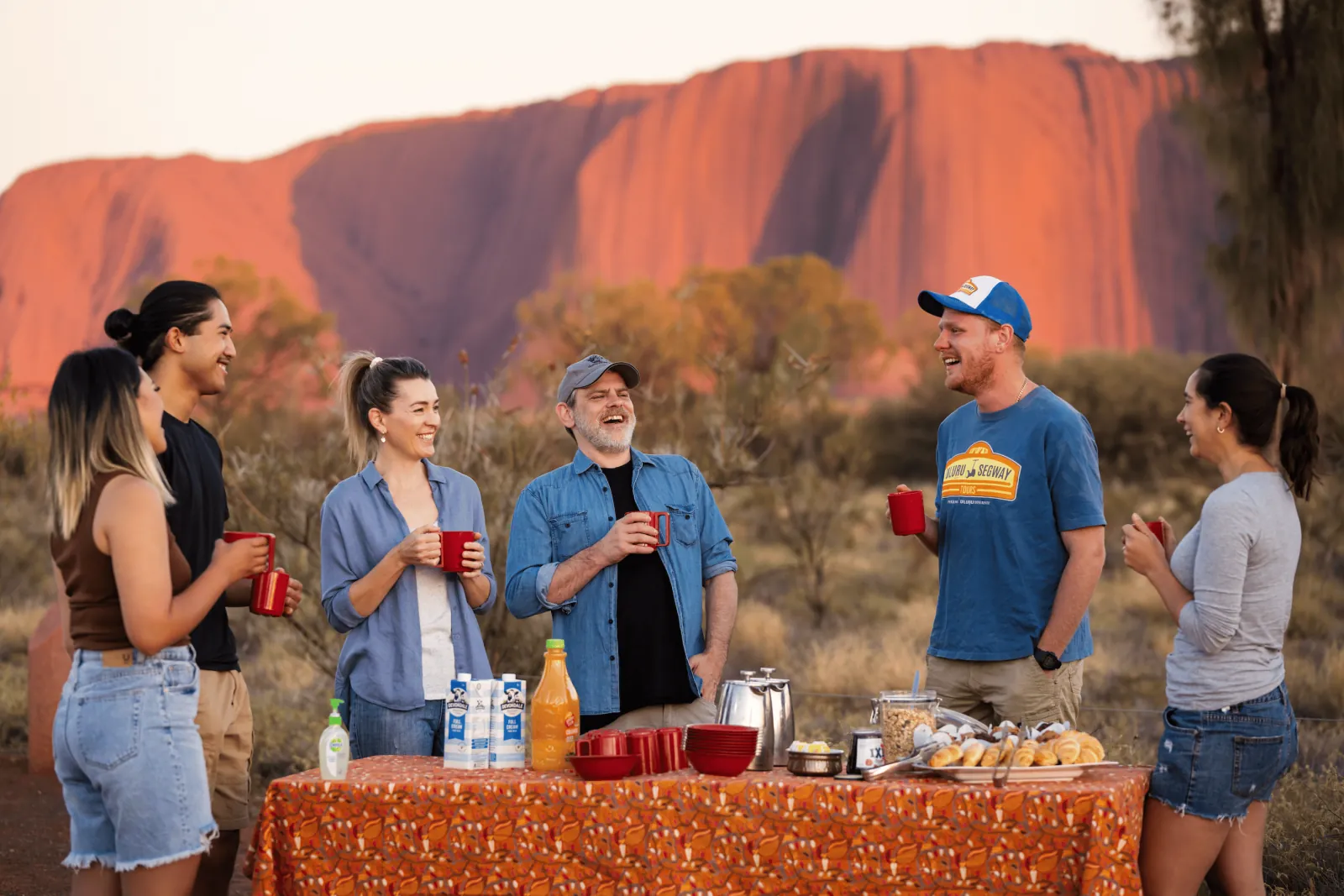 Group with red mugs at a breakfast table of pastries and juice as early light turns Uluru red