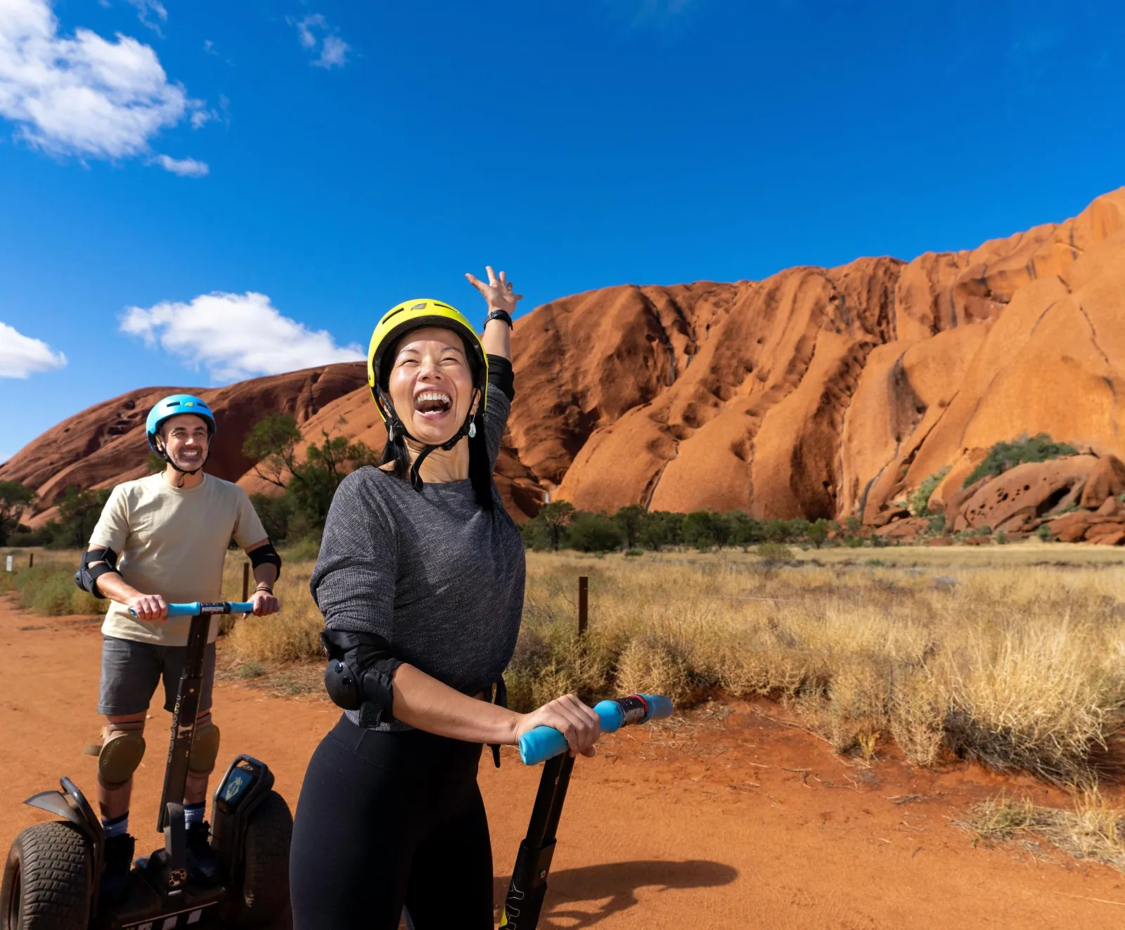Laughing rider in a yellow helmet waving at Uluru's sunlit face with a second rider behind