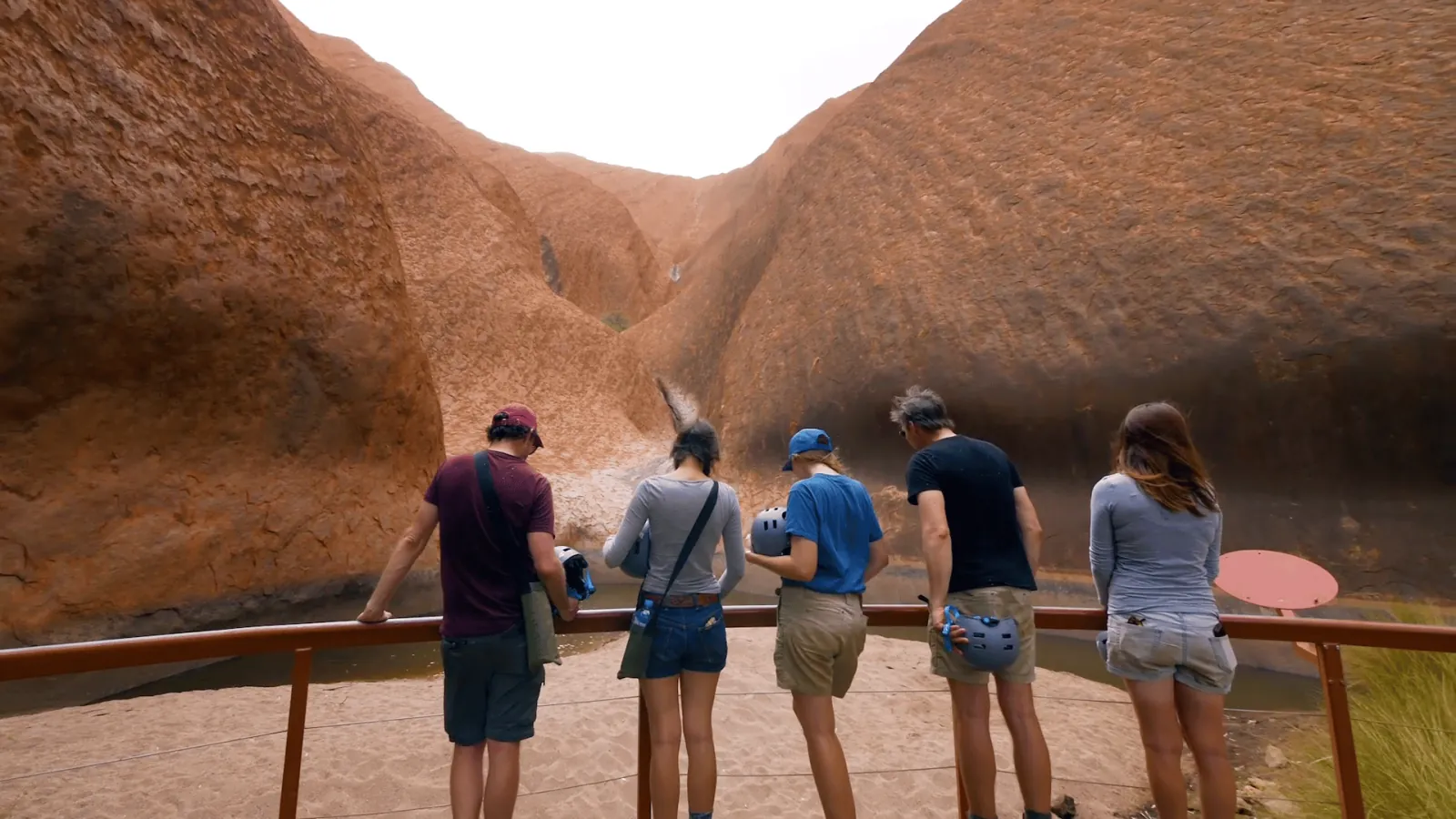 Five visitors holding helmets at a railing above Mutitjulu Waterhole between curved rock walls