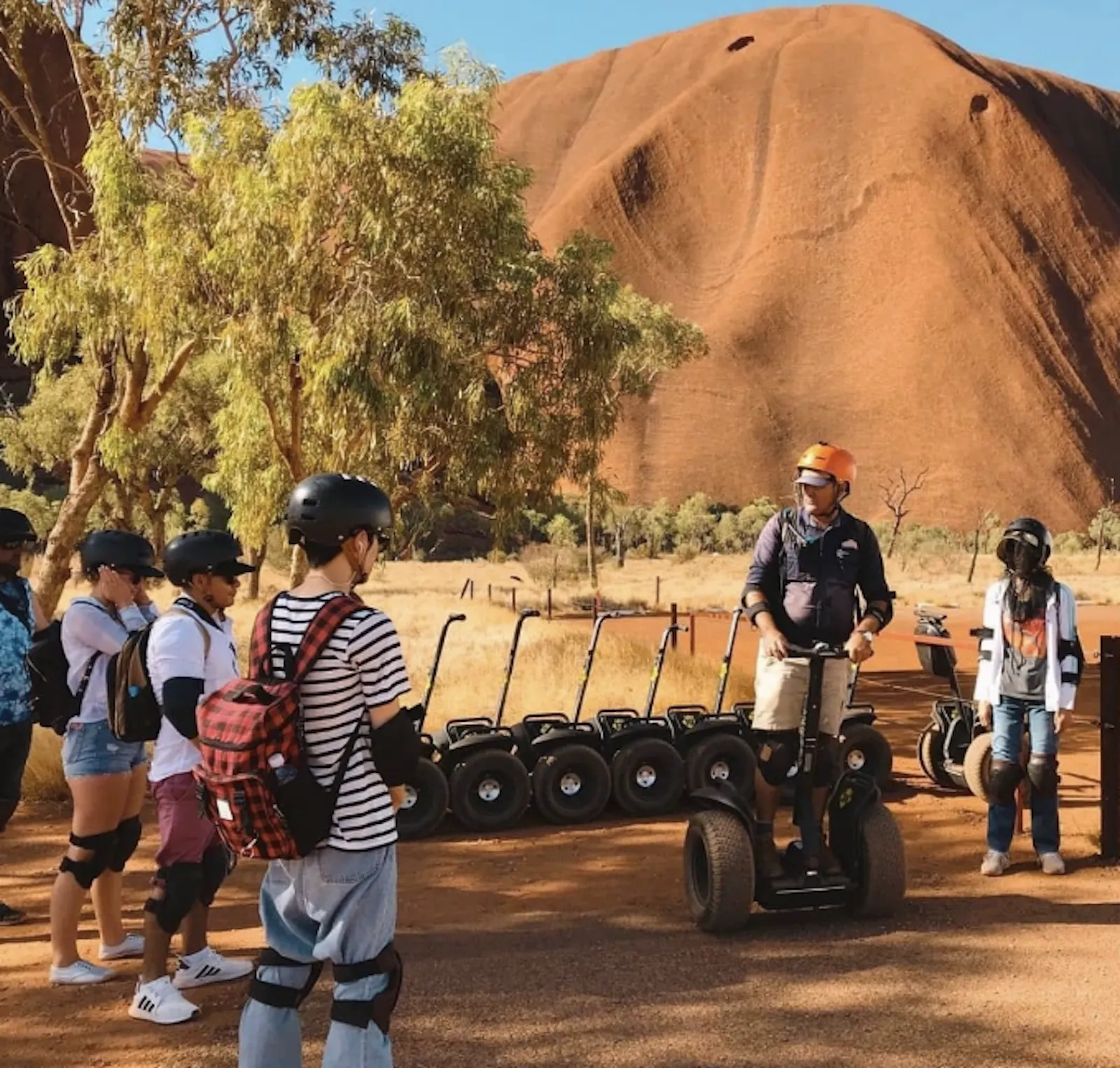 Guide in an orange helmet briefing helmeted riders beside a row of parked Segways near Uluru