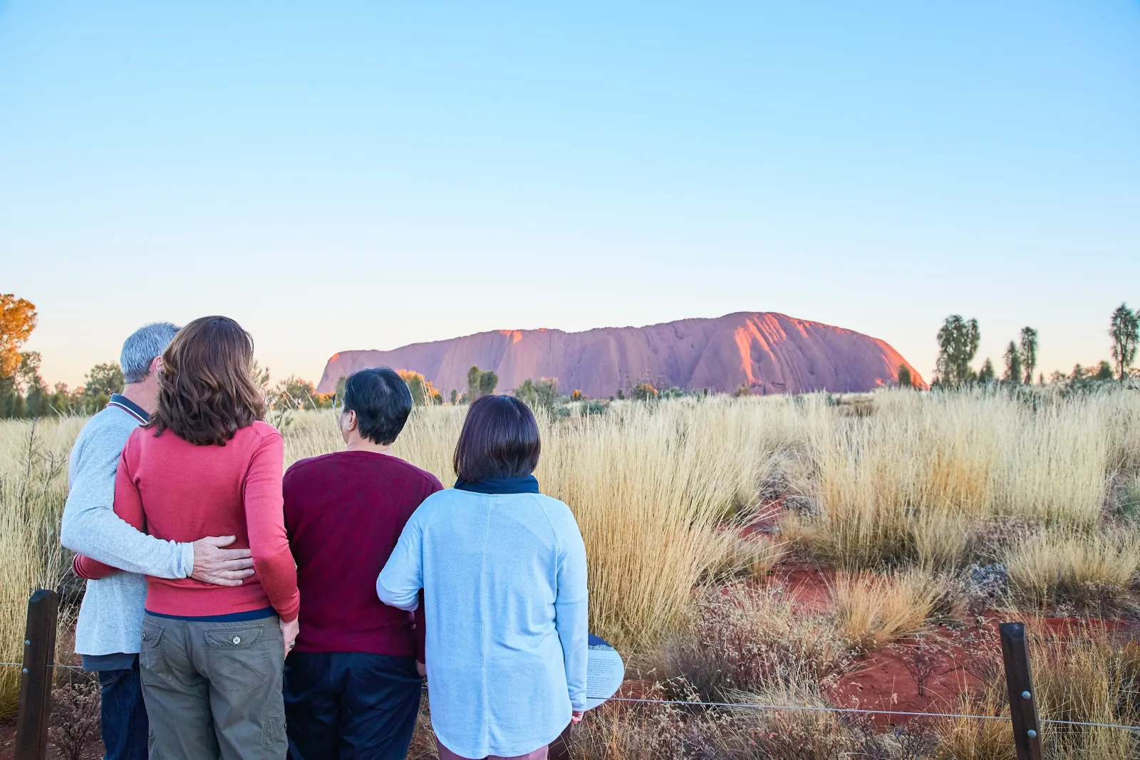 Four visitors in warm layers watching early light turn Uluru pink across spinifex grass