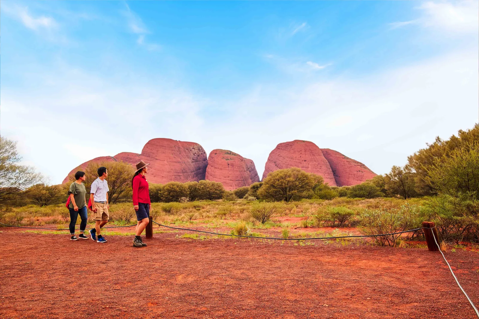 Guide in an Akubra hat leading two walkers along a red gravel path toward the Kata Tjuta domes