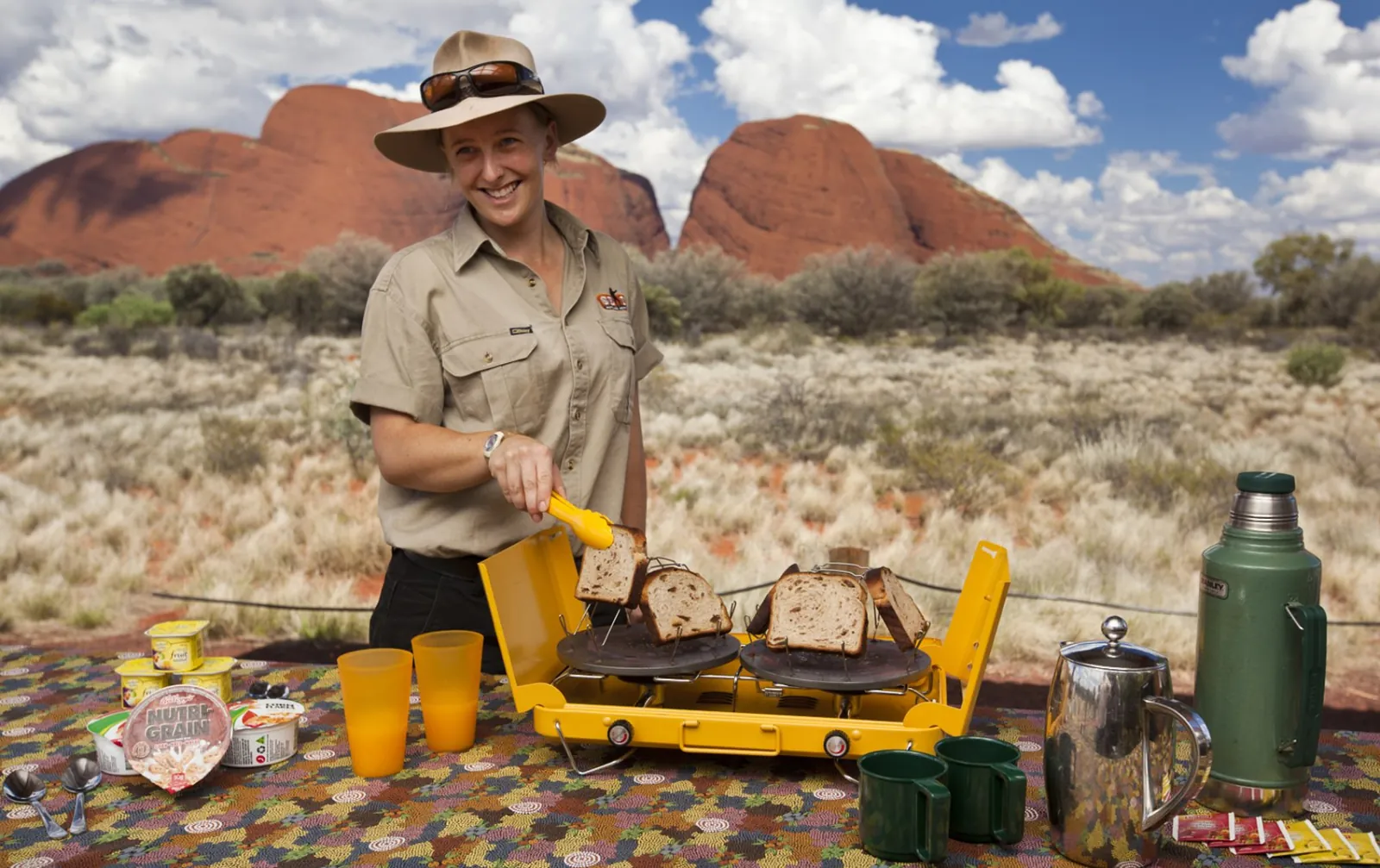Guide toasting bread on a camp stove at a breakfast table with two Kata Tjuta domes behind