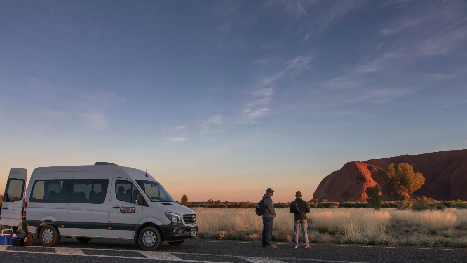 White SEIT minibus parked at a roadside stop as two guests look toward Uluru at first light