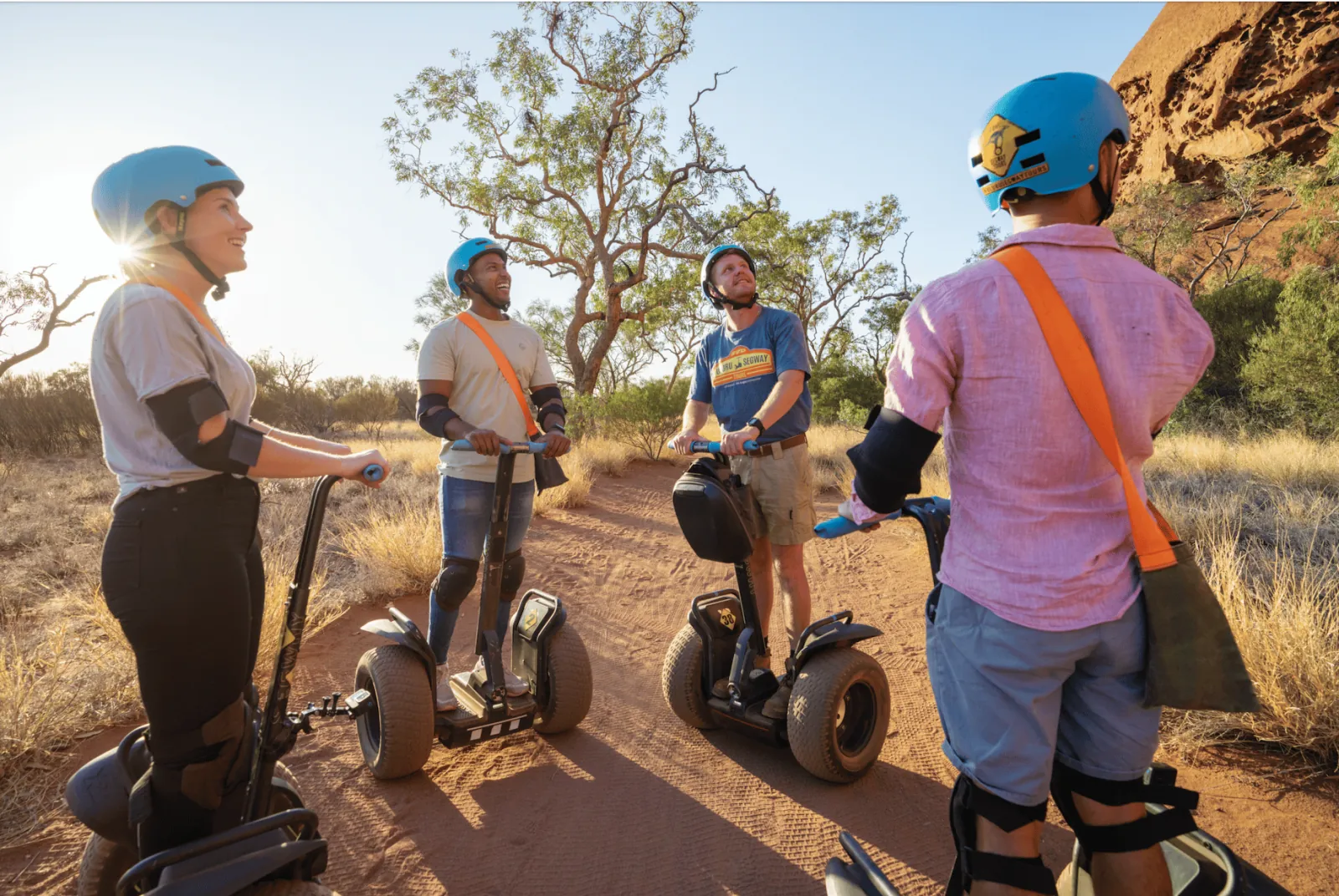 Four Segway riders in helmets and elbow pads chatting on a sandy track beside Uluru