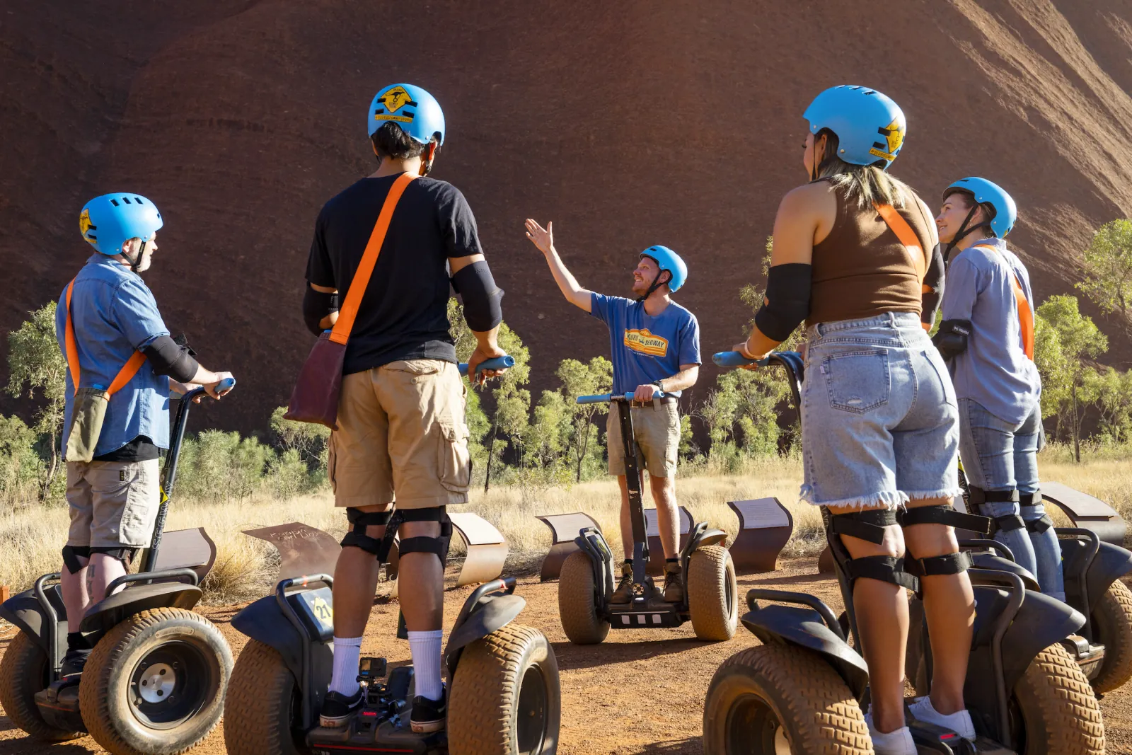 Uluru Segway Tours guide gesturing to the rock while riders in blue helmets listen on their Segways