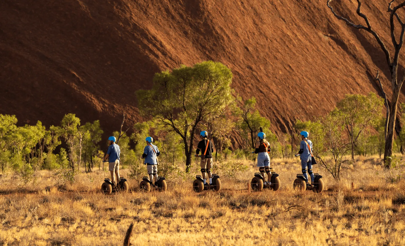 Five Segway riders in a row among desert oaks and dry grass at the foot of Uluru's red wall
