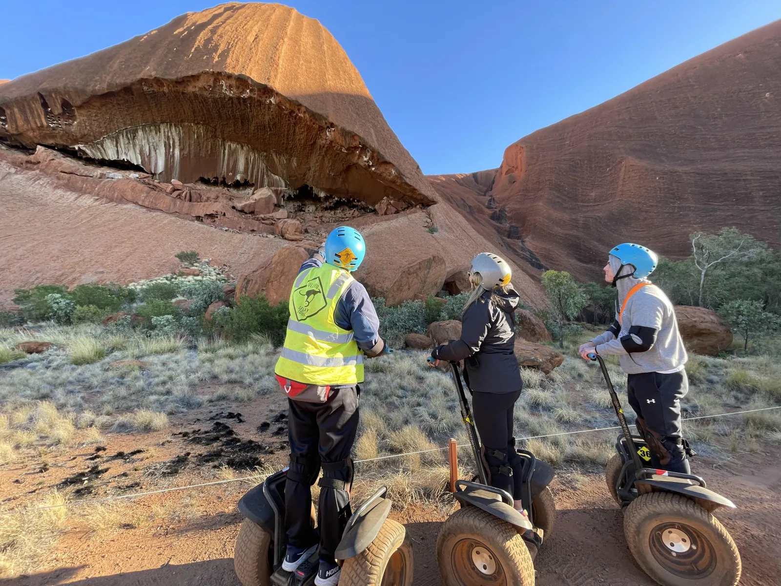 Guide in a hi-vis vest pointing out a deep overhang in Uluru to two riders on Segways