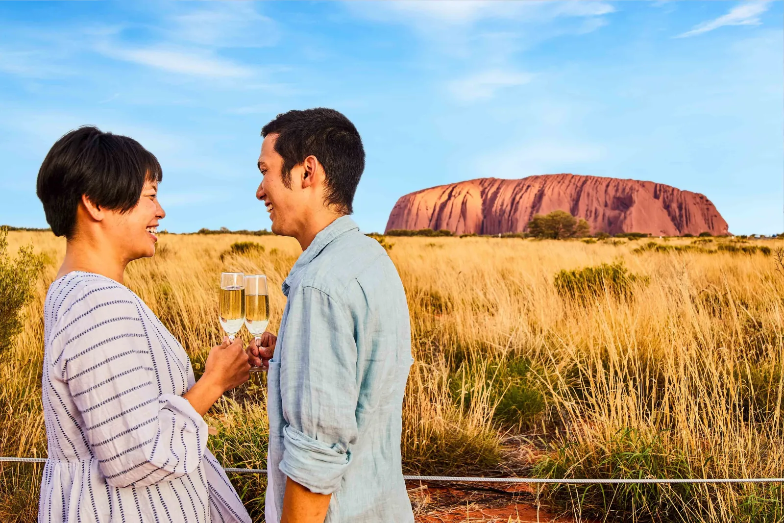 Couple laughing with glasses of sparkling wine at the viewing area fence, Uluru across the grass