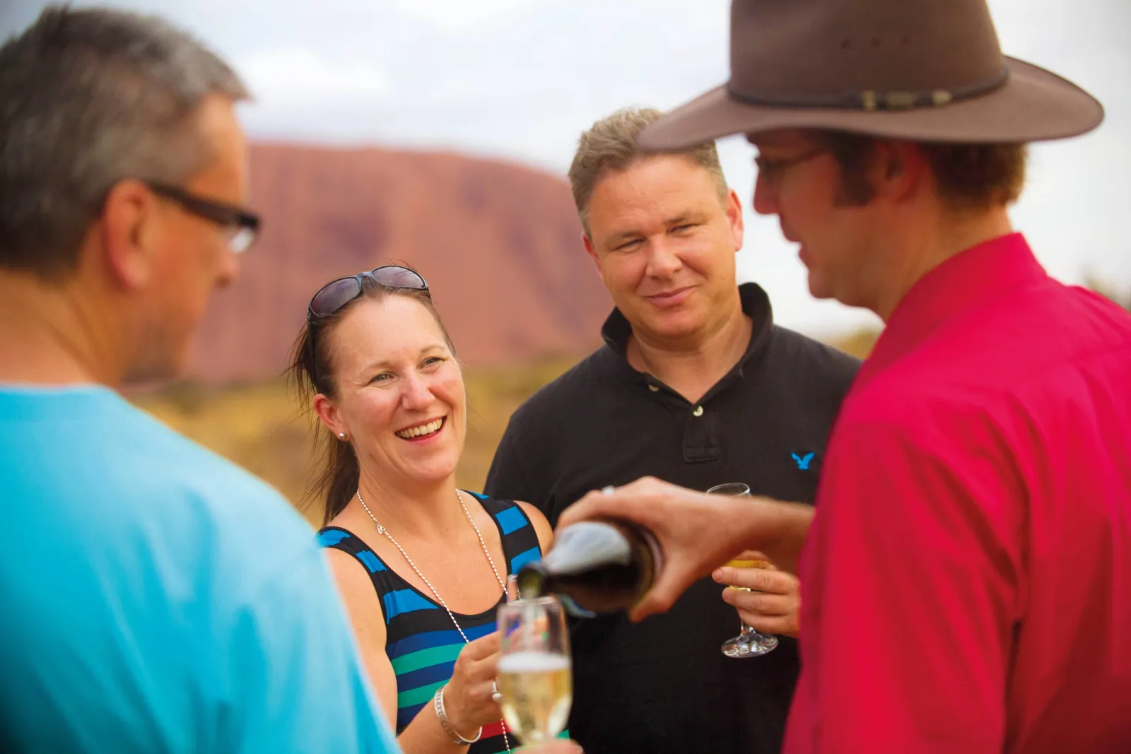 Guide in a bush hat pouring sparkling wine for smiling guests with Uluru blurred behind