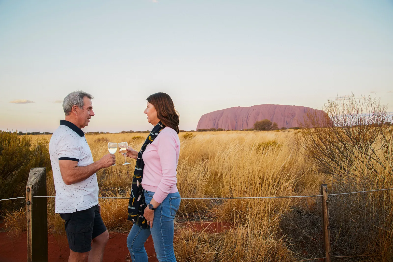 Couple toasting with white wine at the viewing area rail as Uluru turns soft pink at dusk