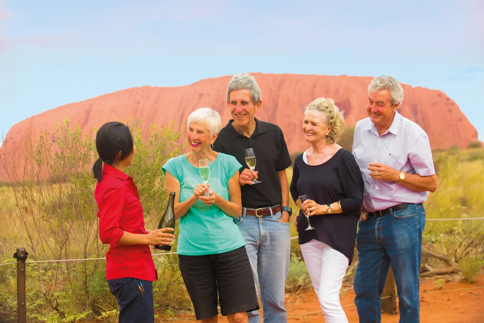 Guide in red holding a bottle as four older travelers chat with wine glasses in front of Uluru