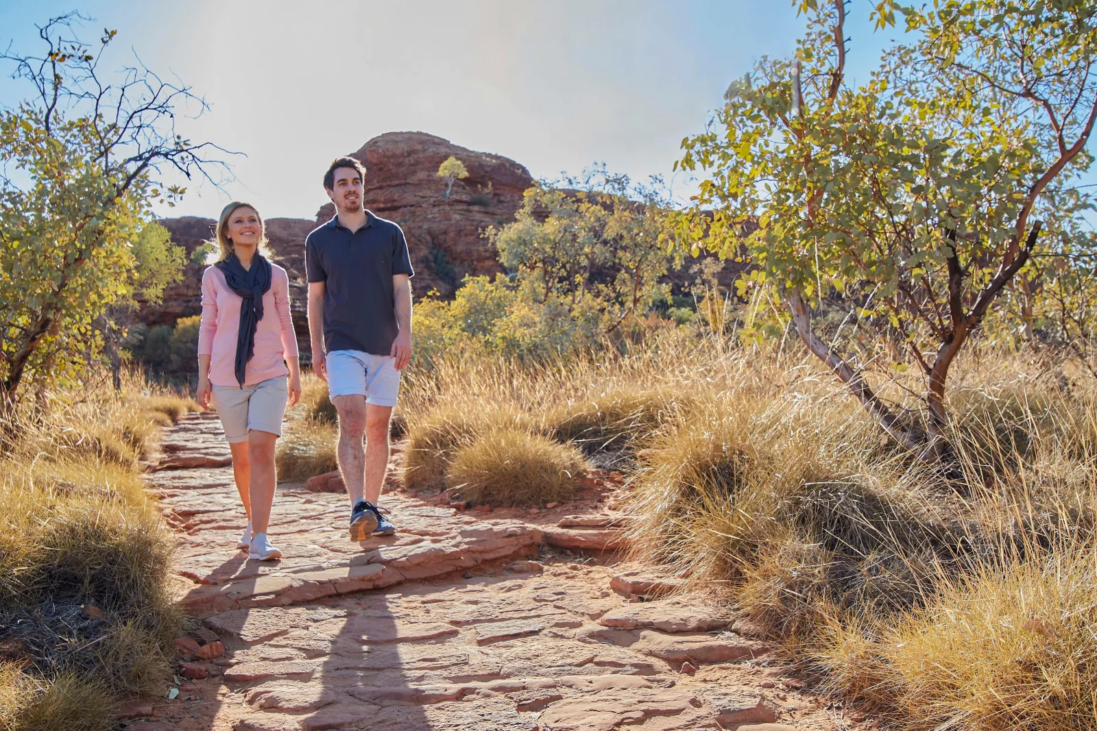 Couple strolling a flat stone path through spinifex and gum trees below red cliffs in morning sun
