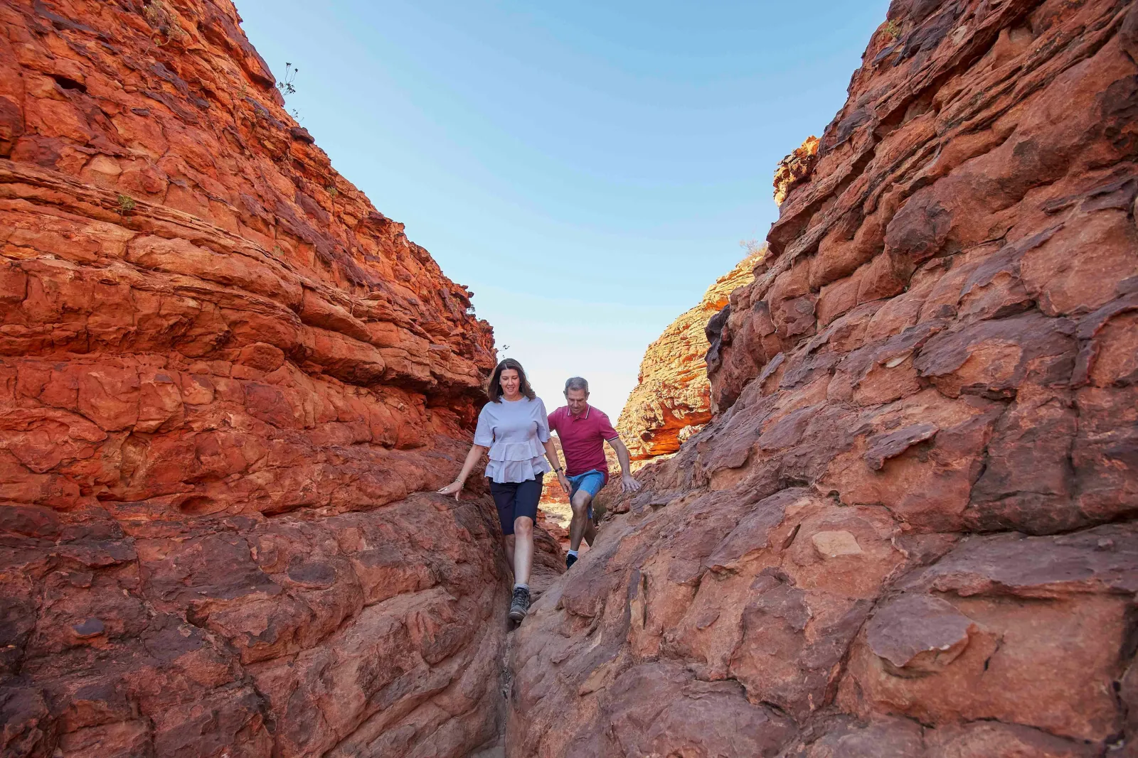 Two hikers stepping down through a narrow cleft between weathered red sandstone walls