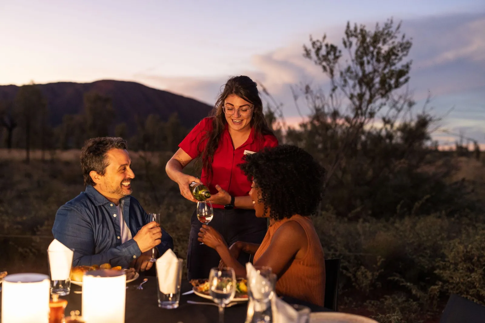 Guide pouring white wine for a couple at a lantern-lit table with Uluru dark behind