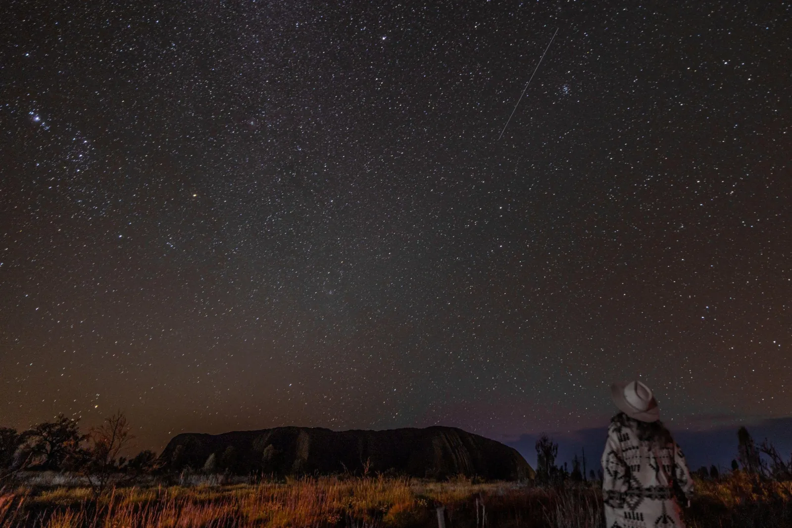 Visitor in a hat gazing at a star-filled sky above the dark outline of Uluru