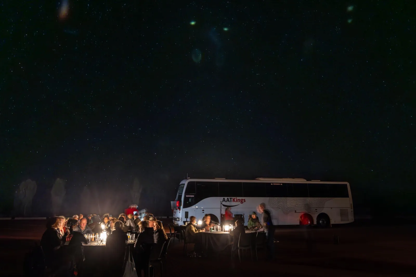 Diners at lantern-lit tables under a starry sky beside a parked AAT Kings coach