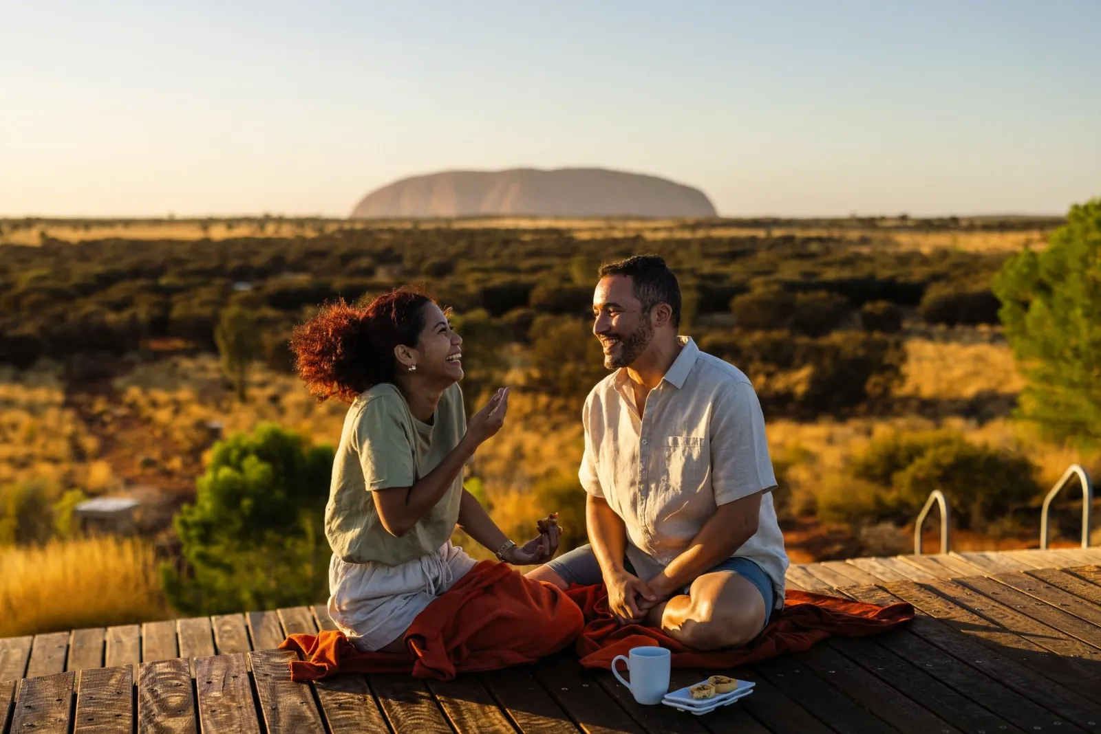 Couple sitting on a wooden deck with a hot drink and pastries, Uluru in soft morning light behind them