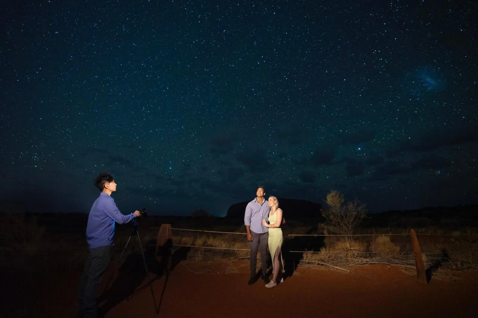 Photographer on a tripod taking a couple's portrait under stars with Uluru dark behind