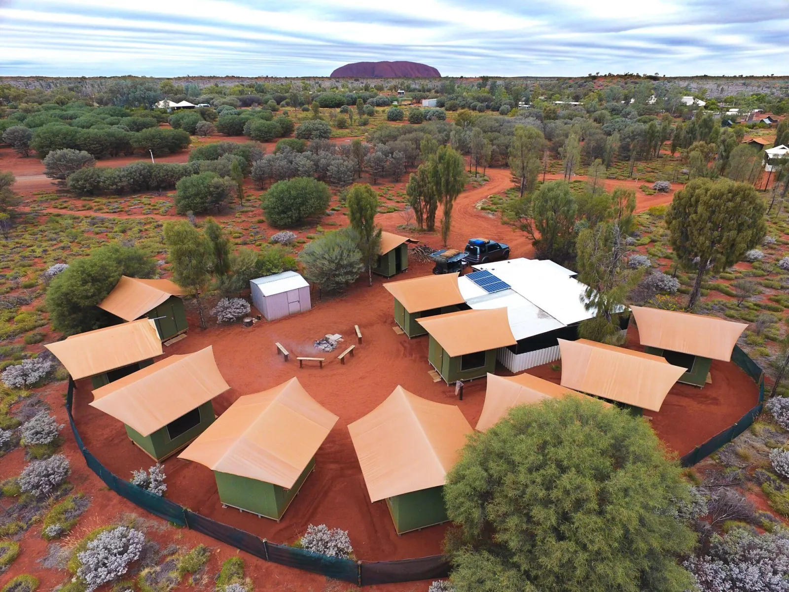 Aerial view of a fenced outback campsite with canvas eco tents around a fire circle and Uluru on the horizon