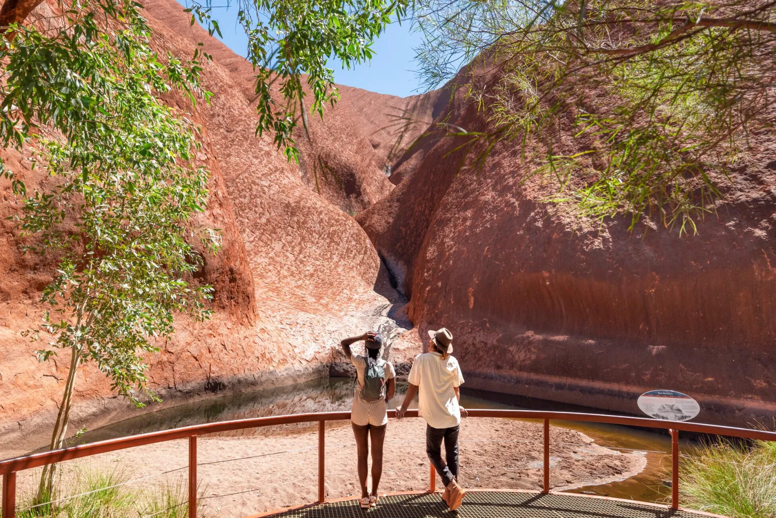 Two visitors at a railed platform looking over the pool at Mutitjulu Waterhole between red rock walls