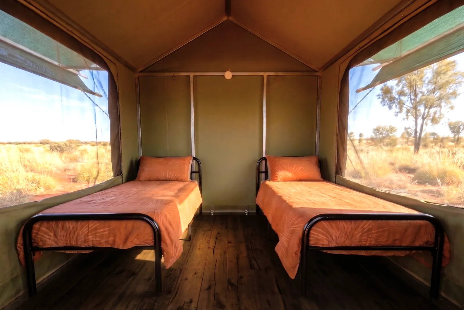 Inside a canvas eco tent: two single beds with orange covers and screened windows onto desert scrub