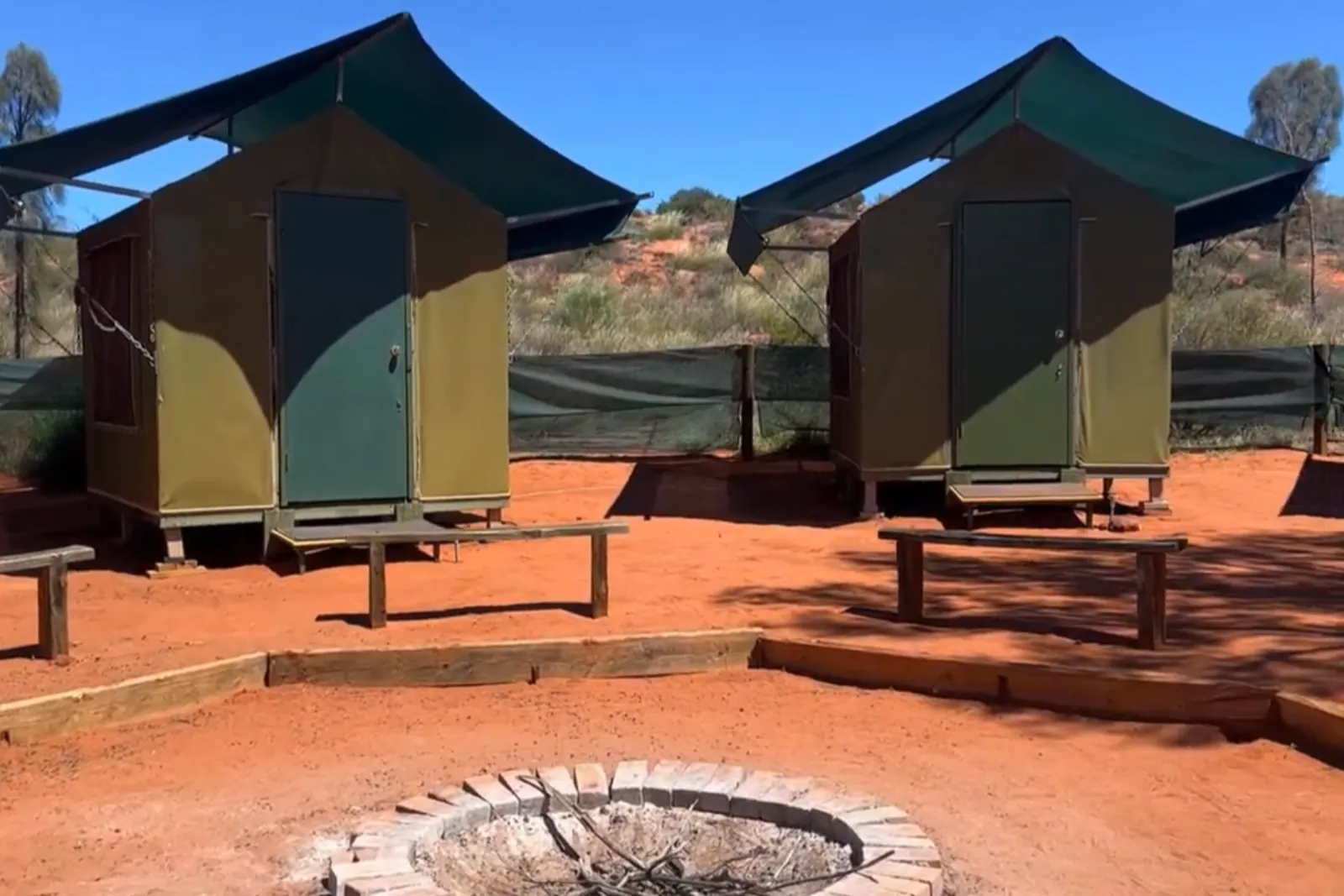 Two green canvas eco tents on raised floors beside benches and a brick fire pit on red sand
