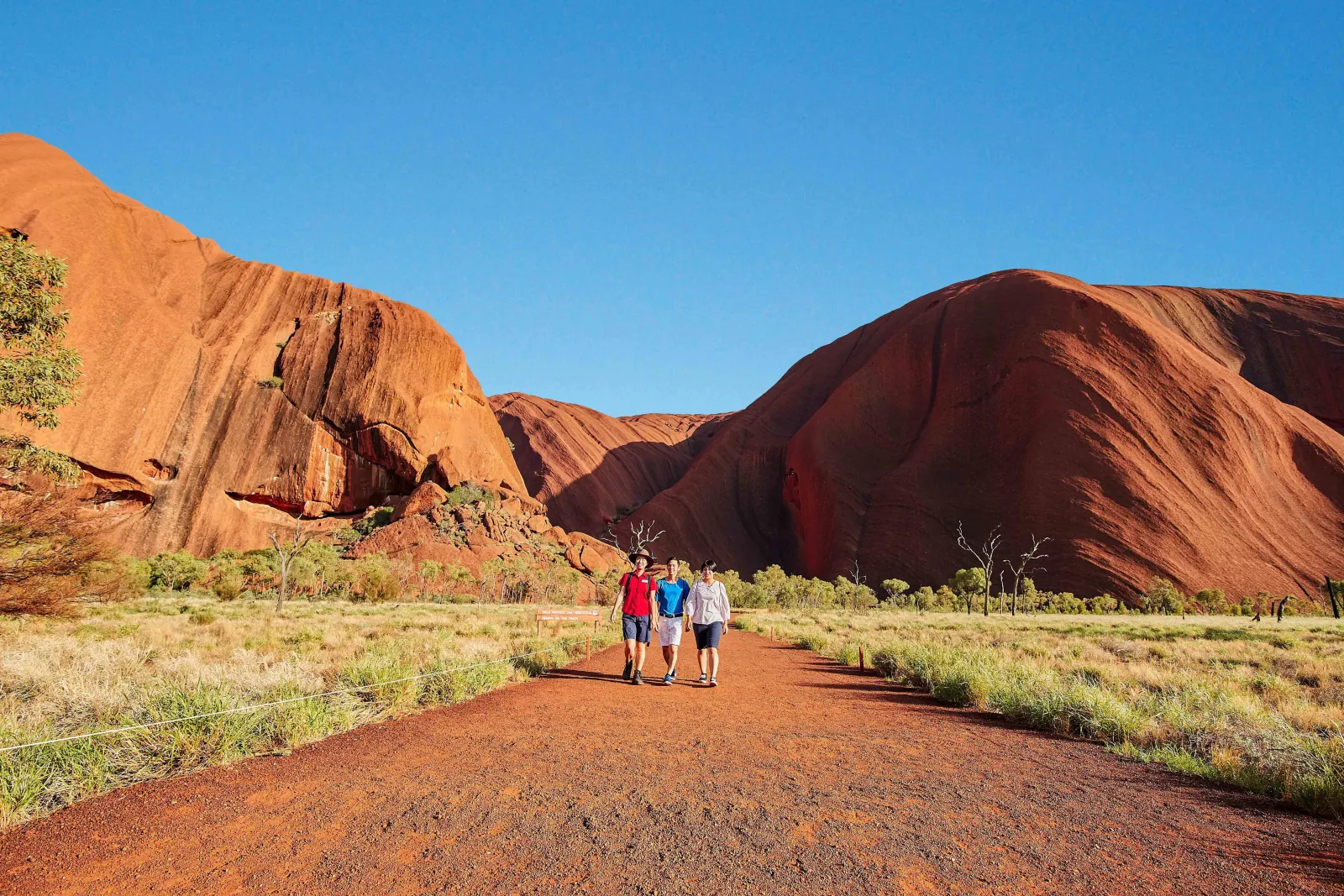 Guide in a red shirt leading two visitors along a red gravel track at the foot of Uluru