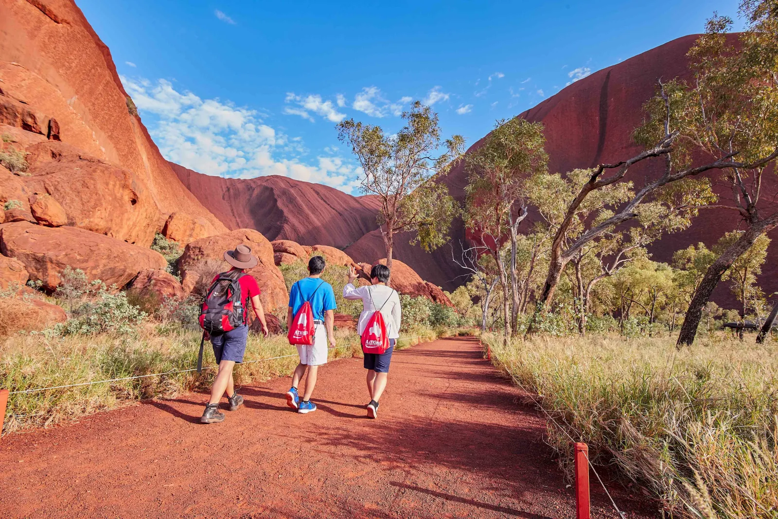Three walkers with AAT Kings bags on a shaded red track between Uluru's cliffs and gum trees