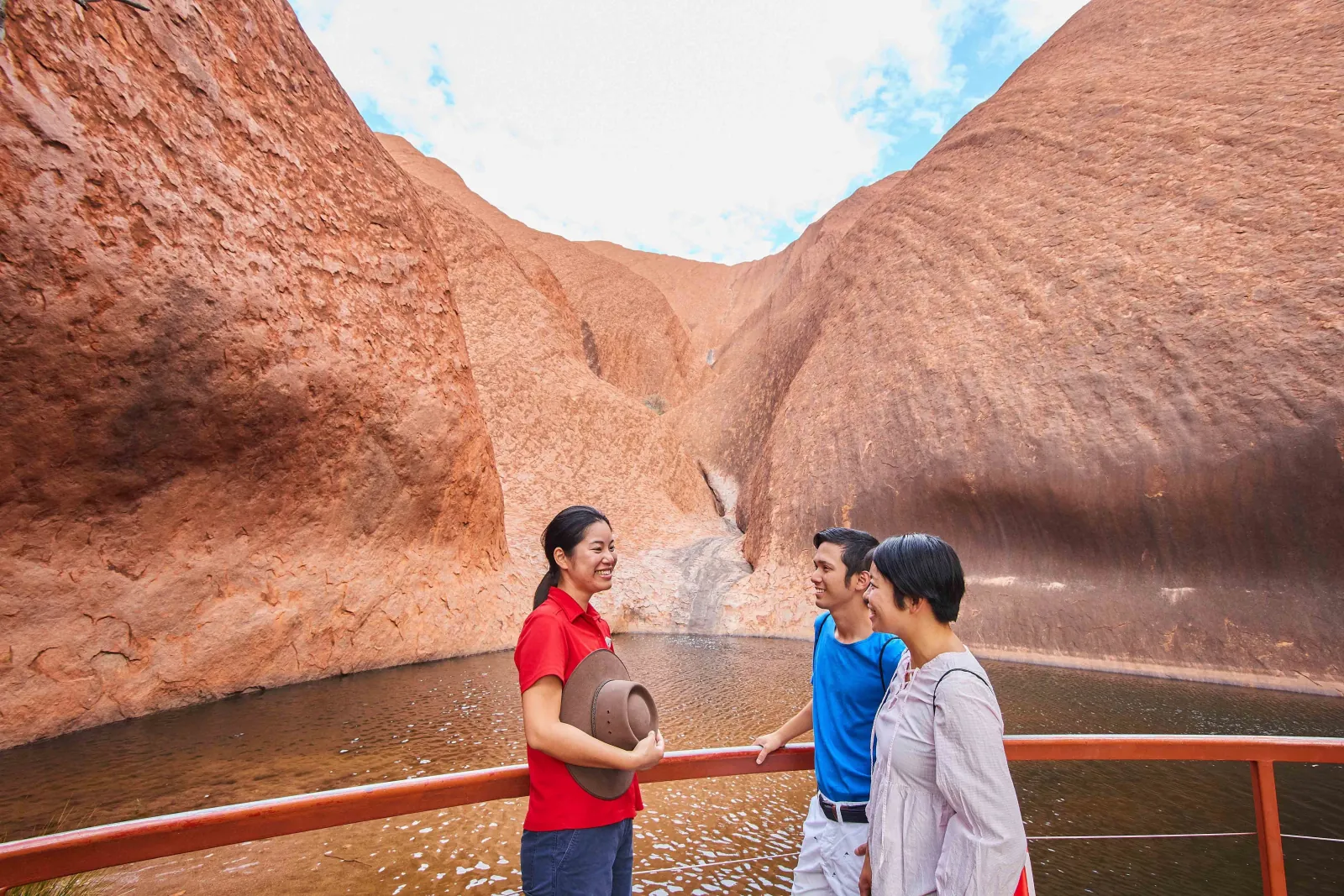 Guide talking with two visitors at the railing above Mutitjulu Waterhole under curved rock walls