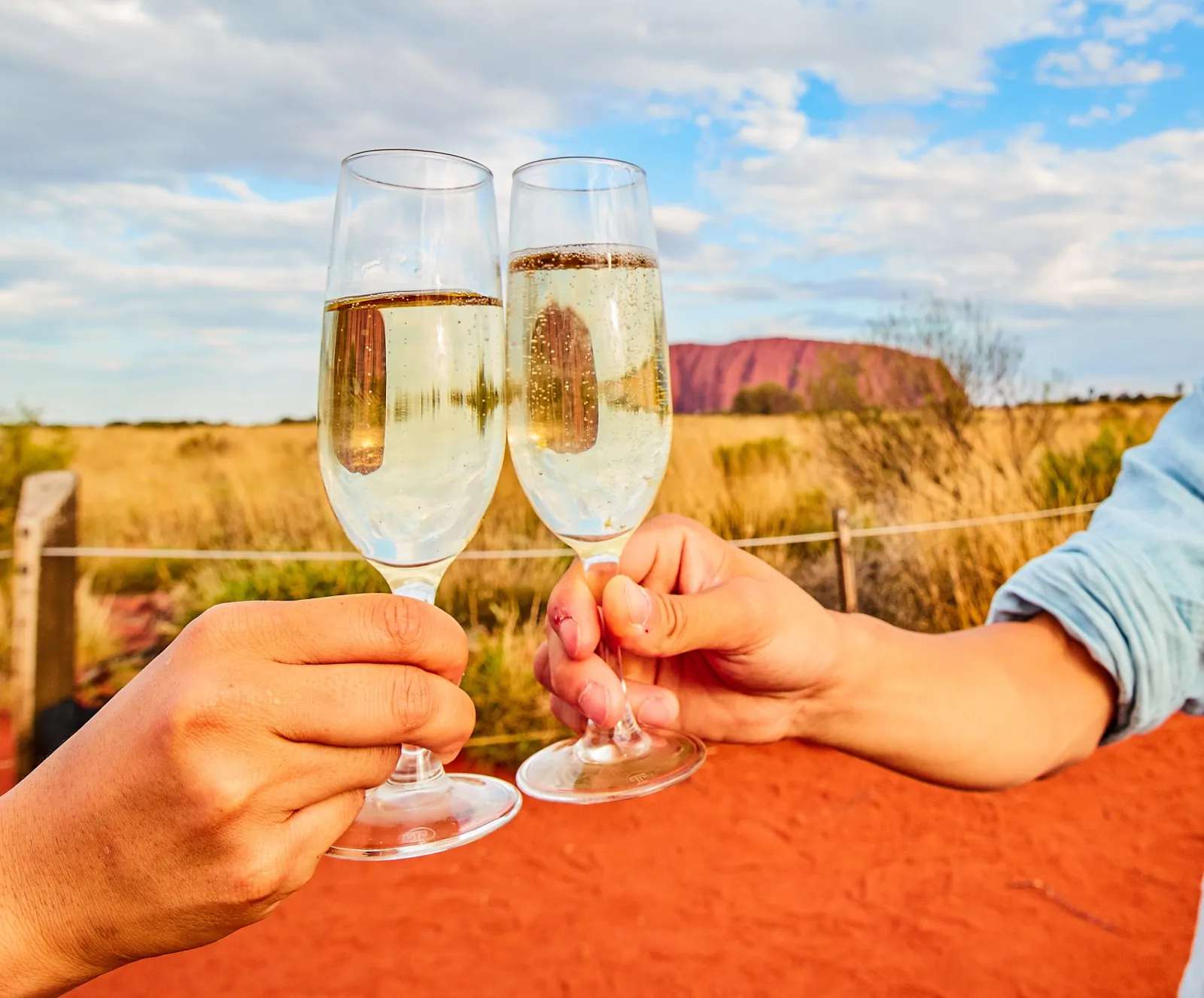 Two glasses of sparkling wine raised in a toast with Uluru reflected in the glass and behind