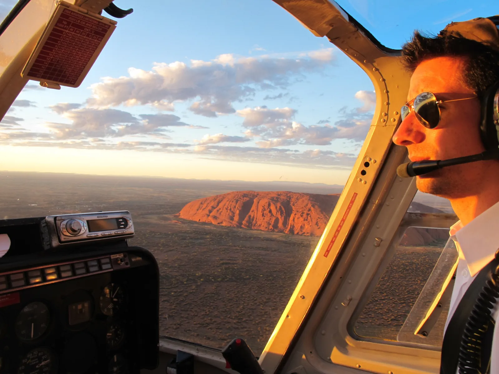 Pilot with headset and sunglasses at the controls, Uluru glowing red through the cockpit window