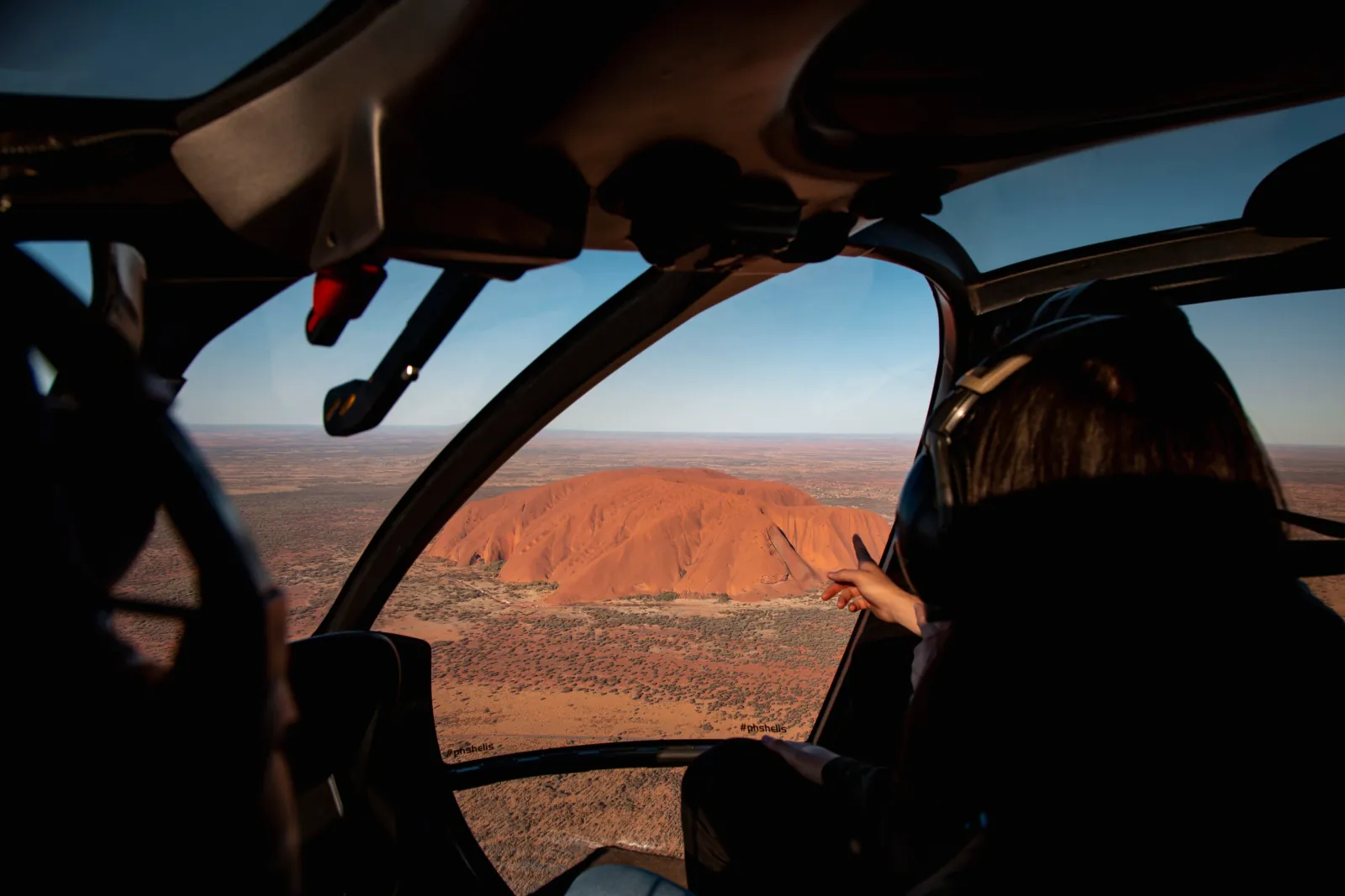 Passenger in a helicopter cabin pointing through the canopy at Uluru on the desert plain below