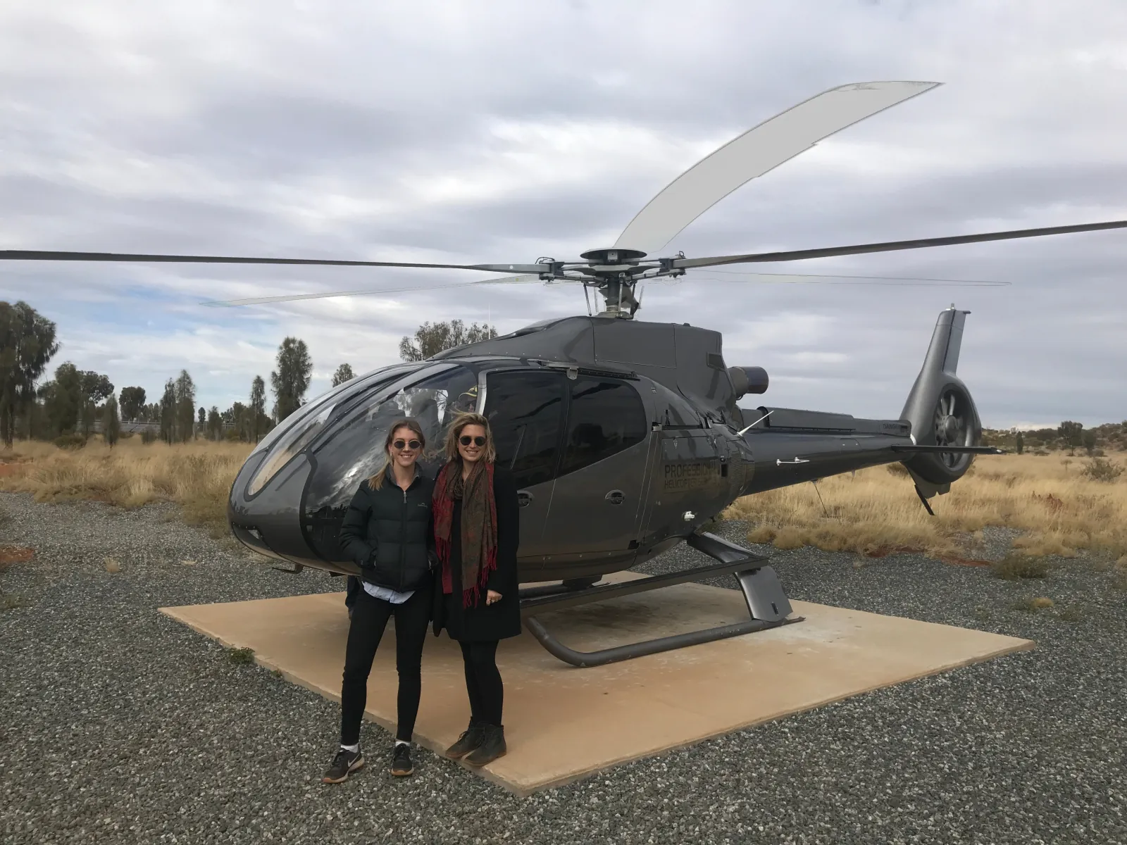 Two passengers in winter jackets standing beside a dark grey helicopter on a gravel helipad