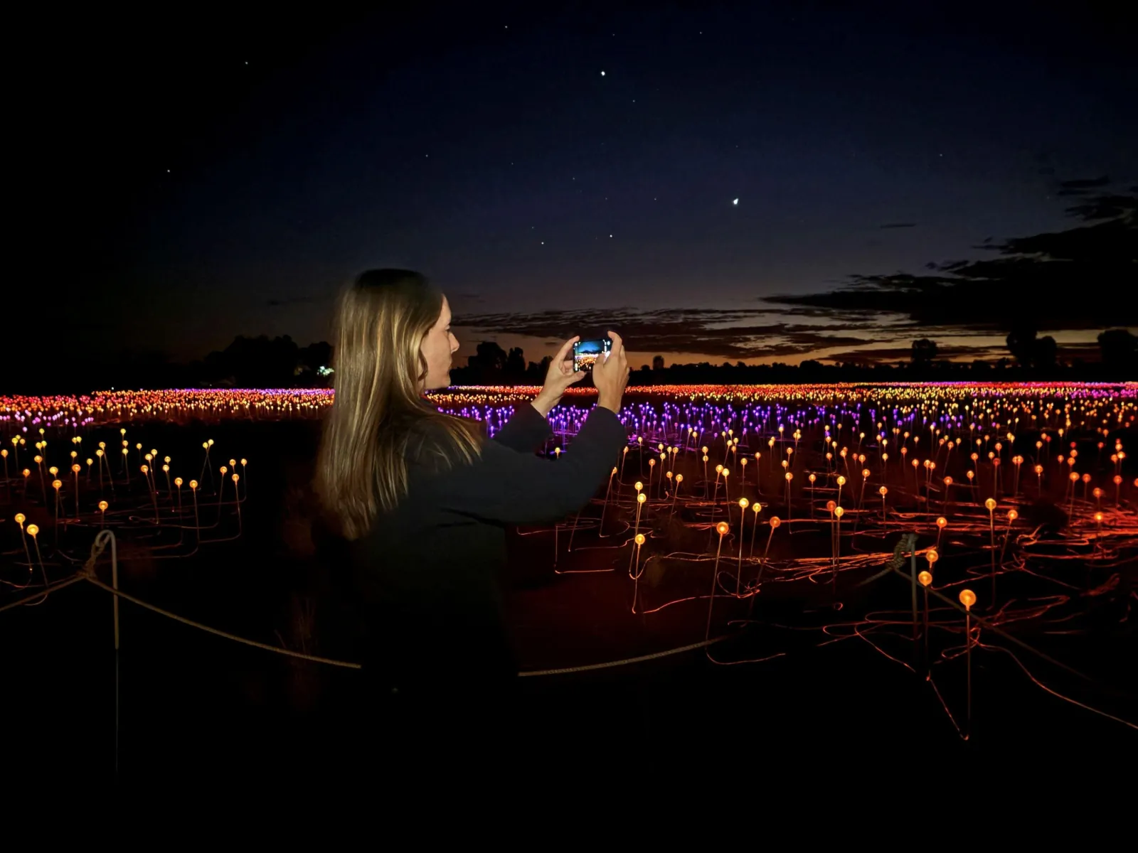 Visitor photographing rows of orange and purple Field of Light stems at first light