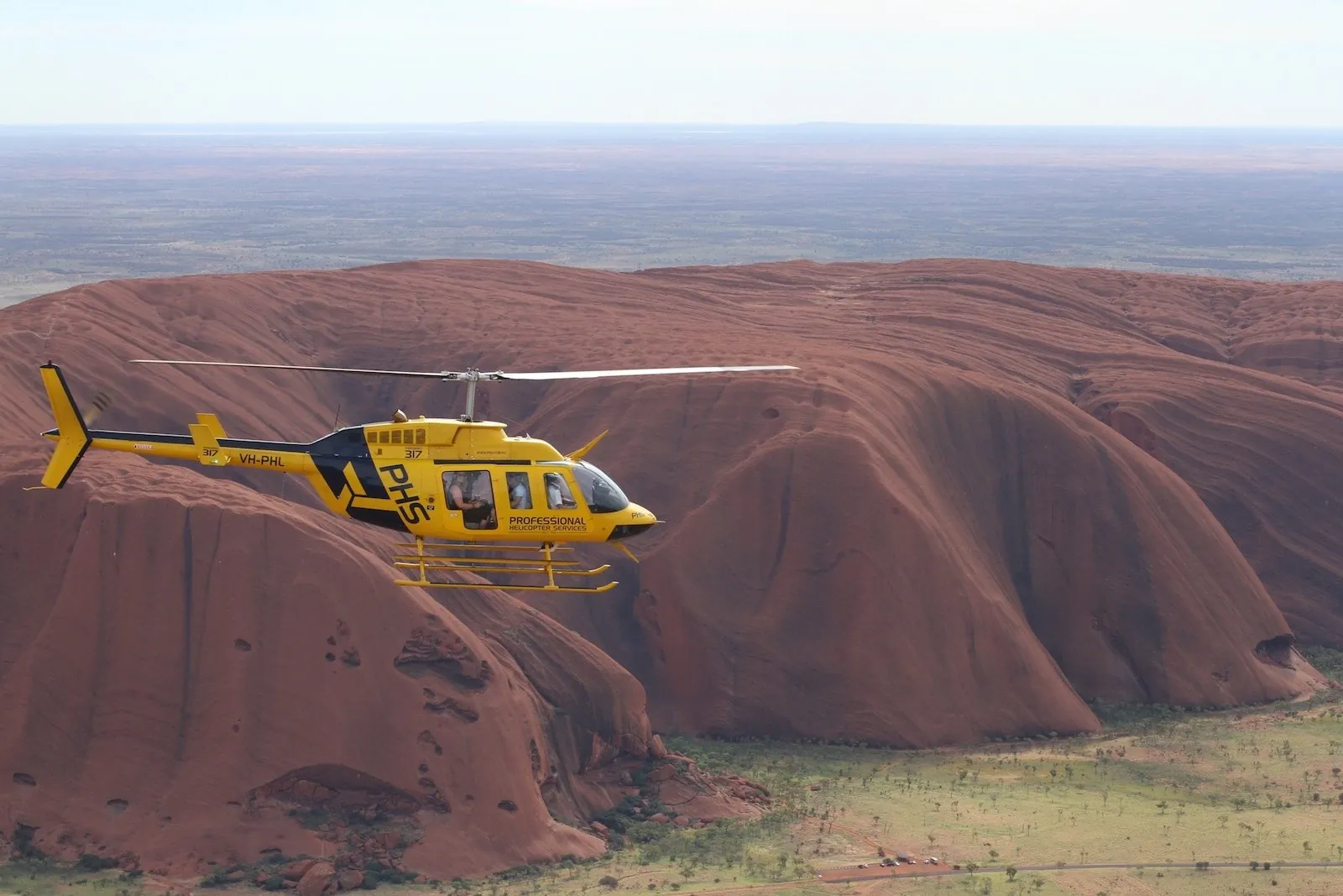 Close view of a yellow Professional Helicopter Services helicopter passing Uluru's ribbed red flank
