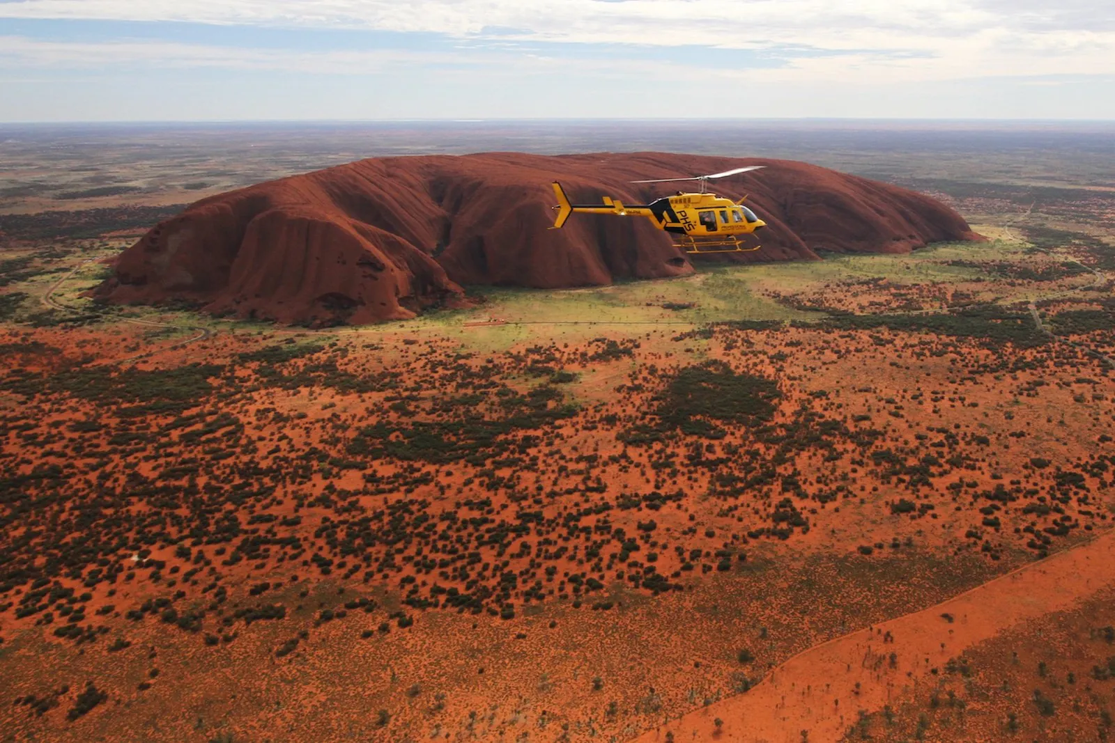 Yellow PHS helicopter flying alongside Uluru with red sand and green scrub stretching to the horizon