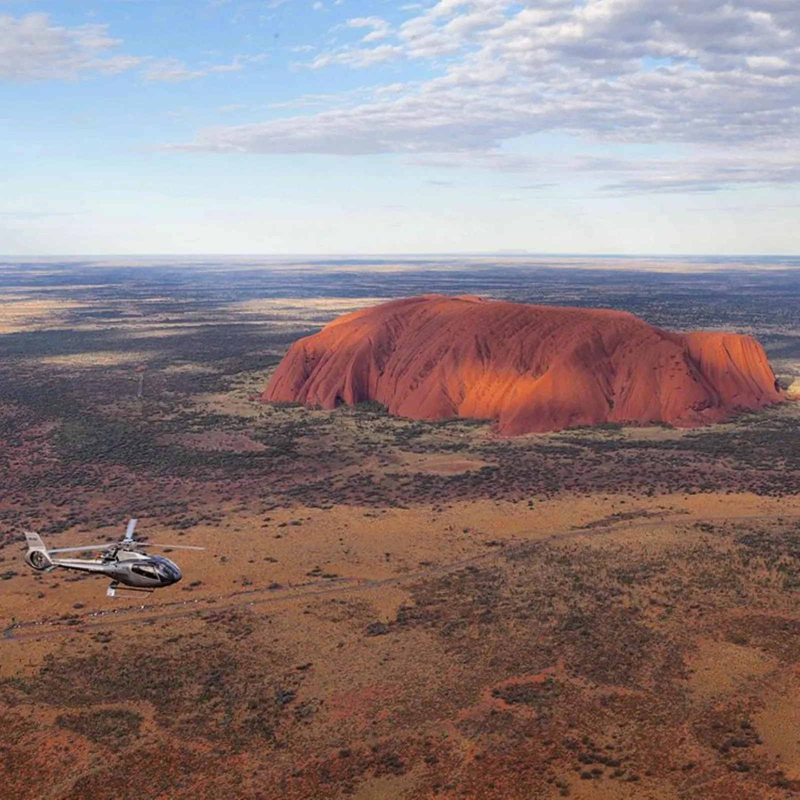 Silver helicopter flying over the desert plain with Uluru lit red in the distance under scattered cloud