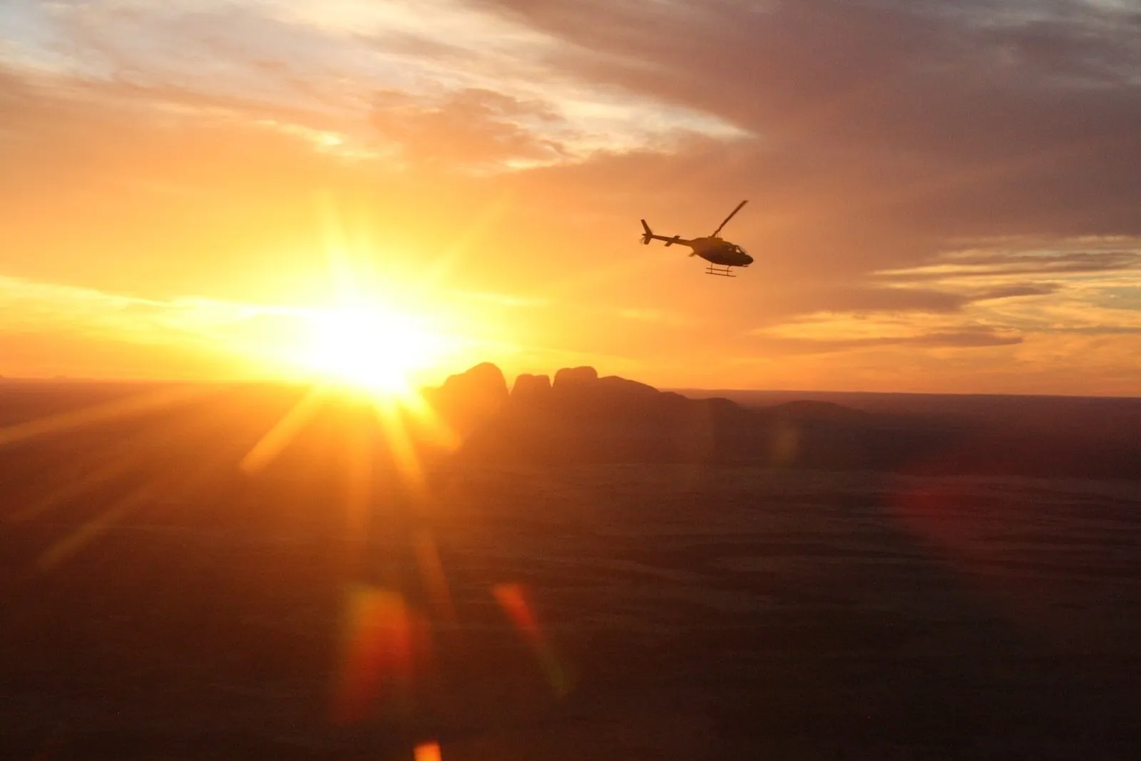 Helicopter silhouetted against a bright sunset with the domes of Kata Tjuta dark on the horizon