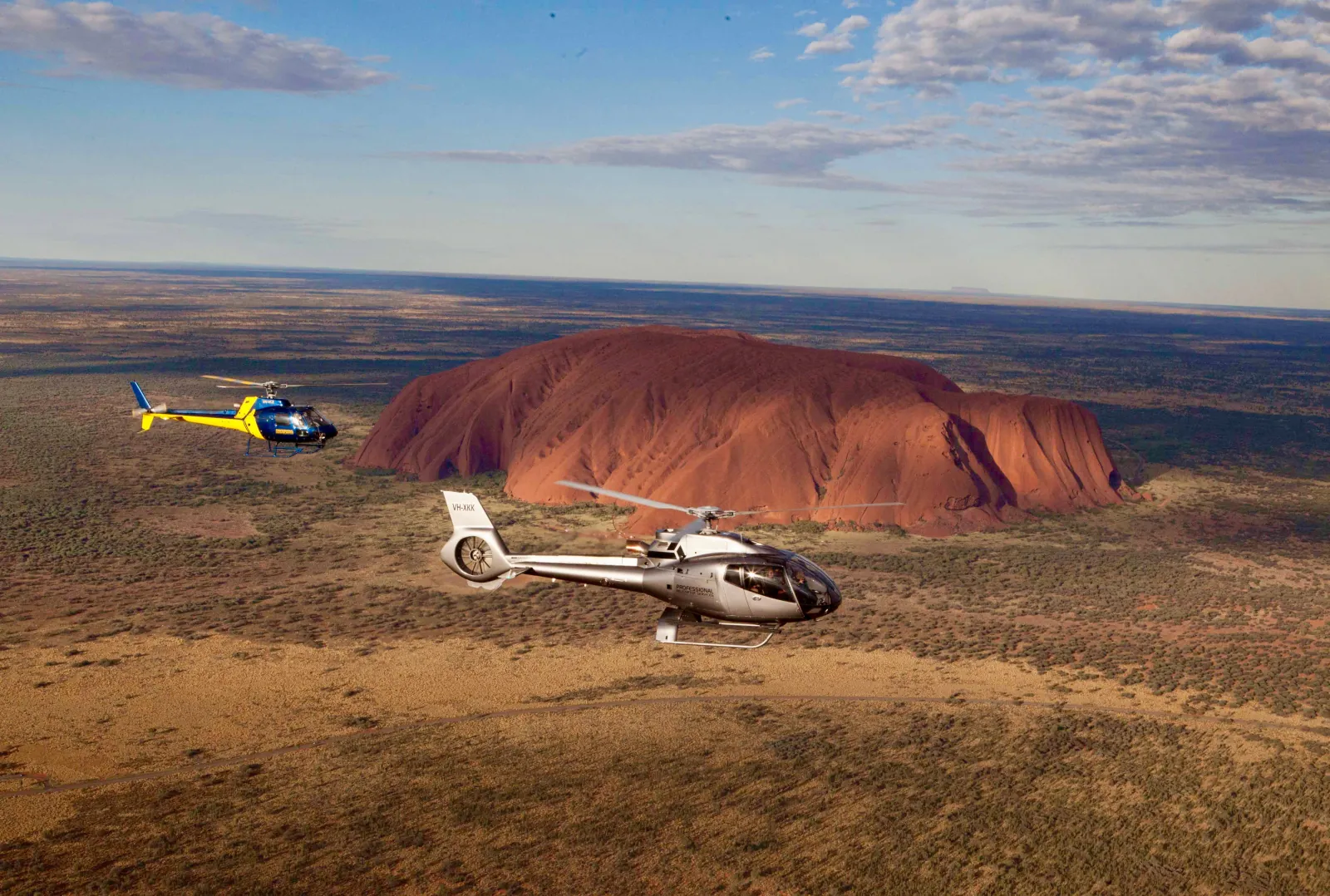 Silver and blue-yellow helicopters flying in front of Uluru across a wide plain under scattered cloud