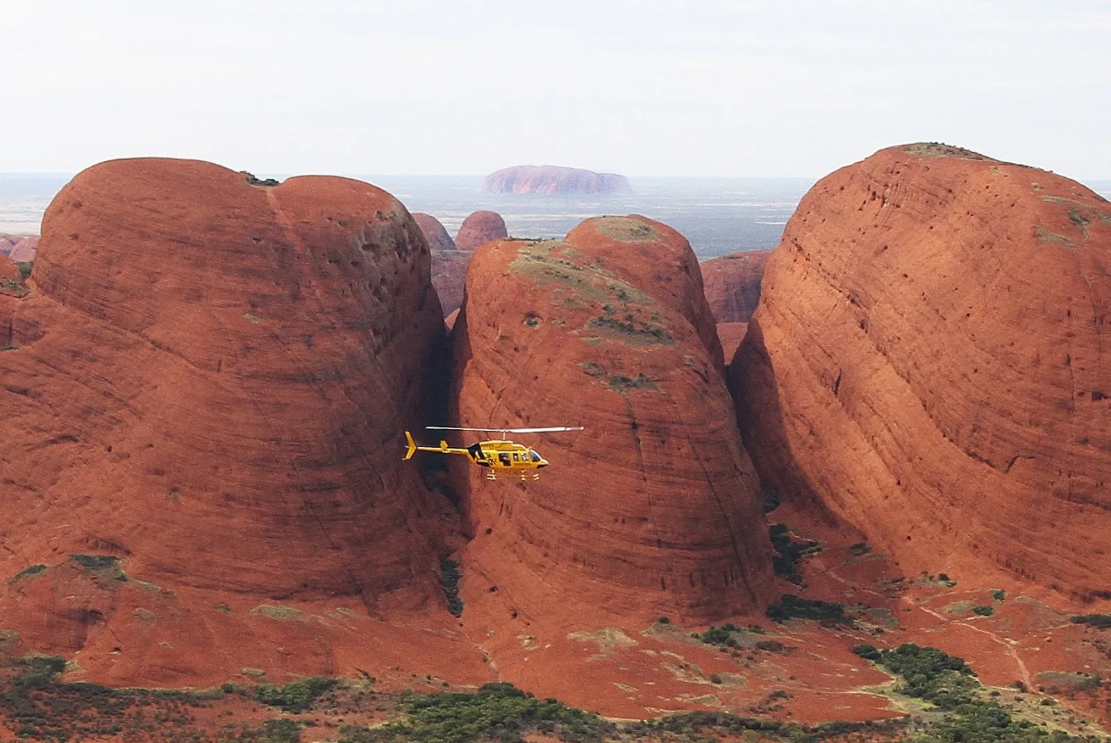 Yellow helicopter dwarfed by three steep Kata Tjuta domes, with Uluru visible far beyond the gap