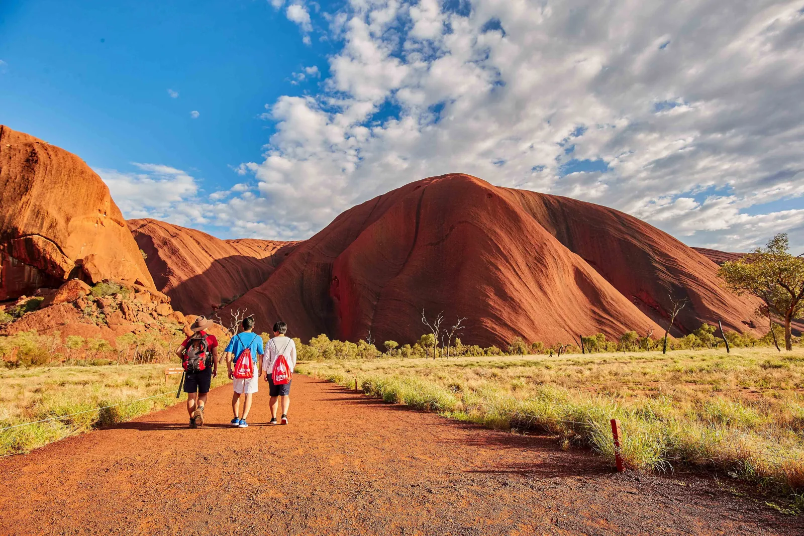 Guide with a backpack leading two walkers along the red base track beside a rounded wall of Uluru