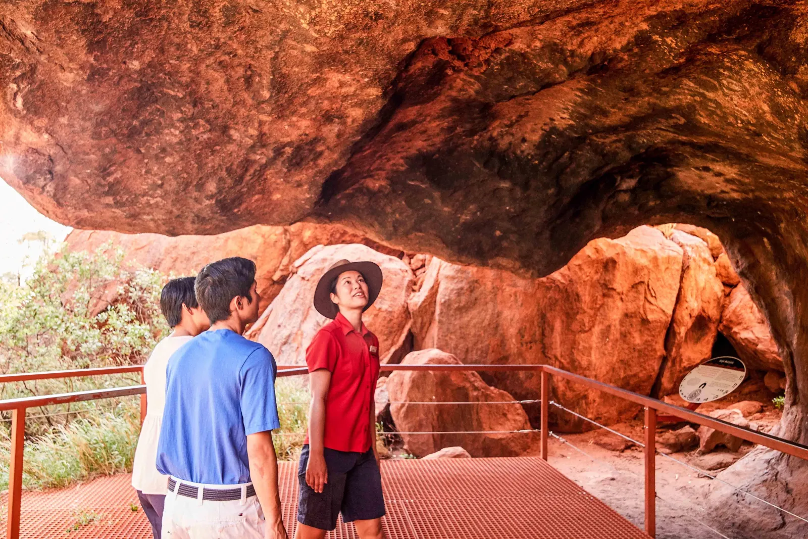Guide in a red shirt and hat showing two guests a rock shelter from a viewing platform