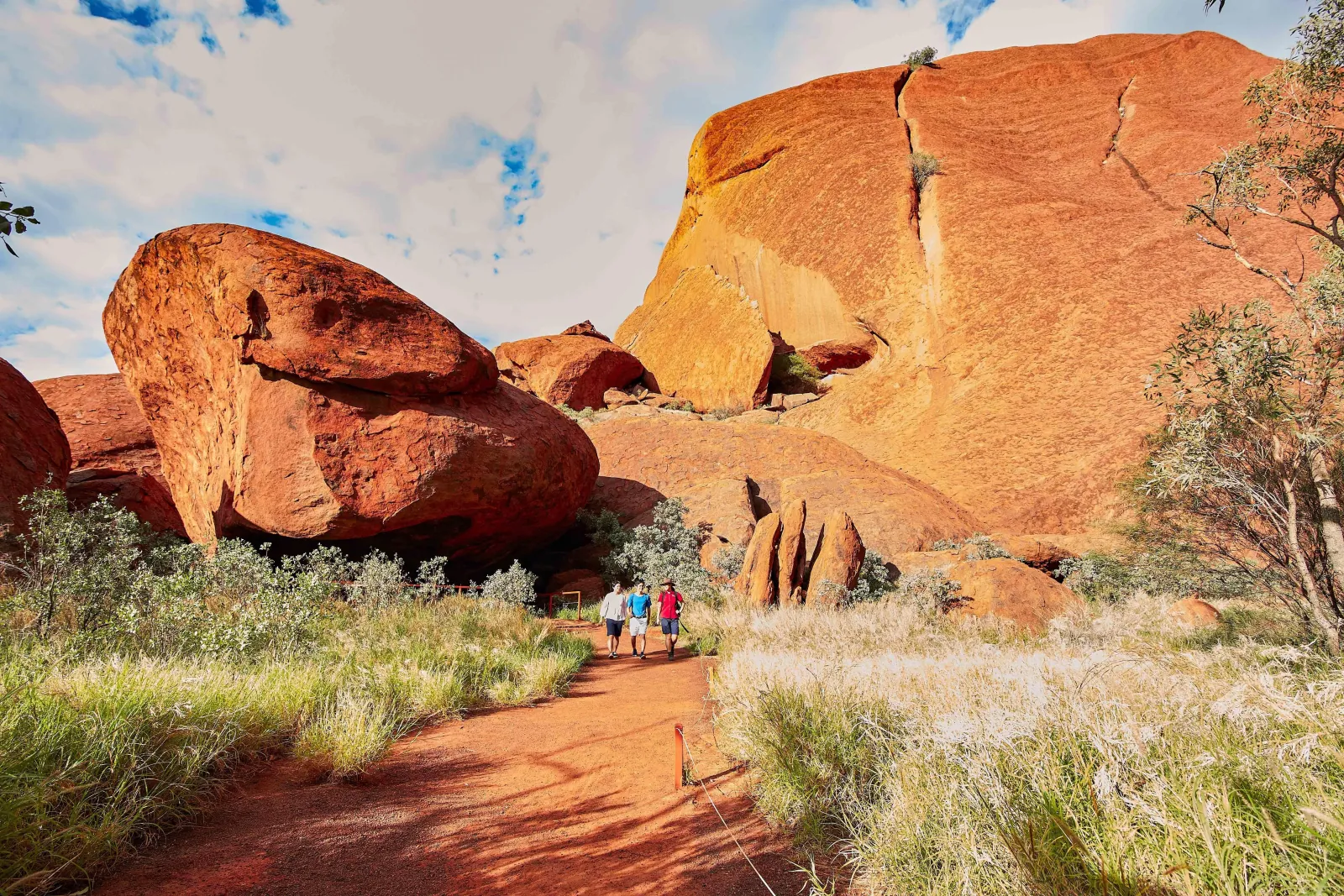 Three walkers on a path among large red boulders and pale grass at the foot of Uluru