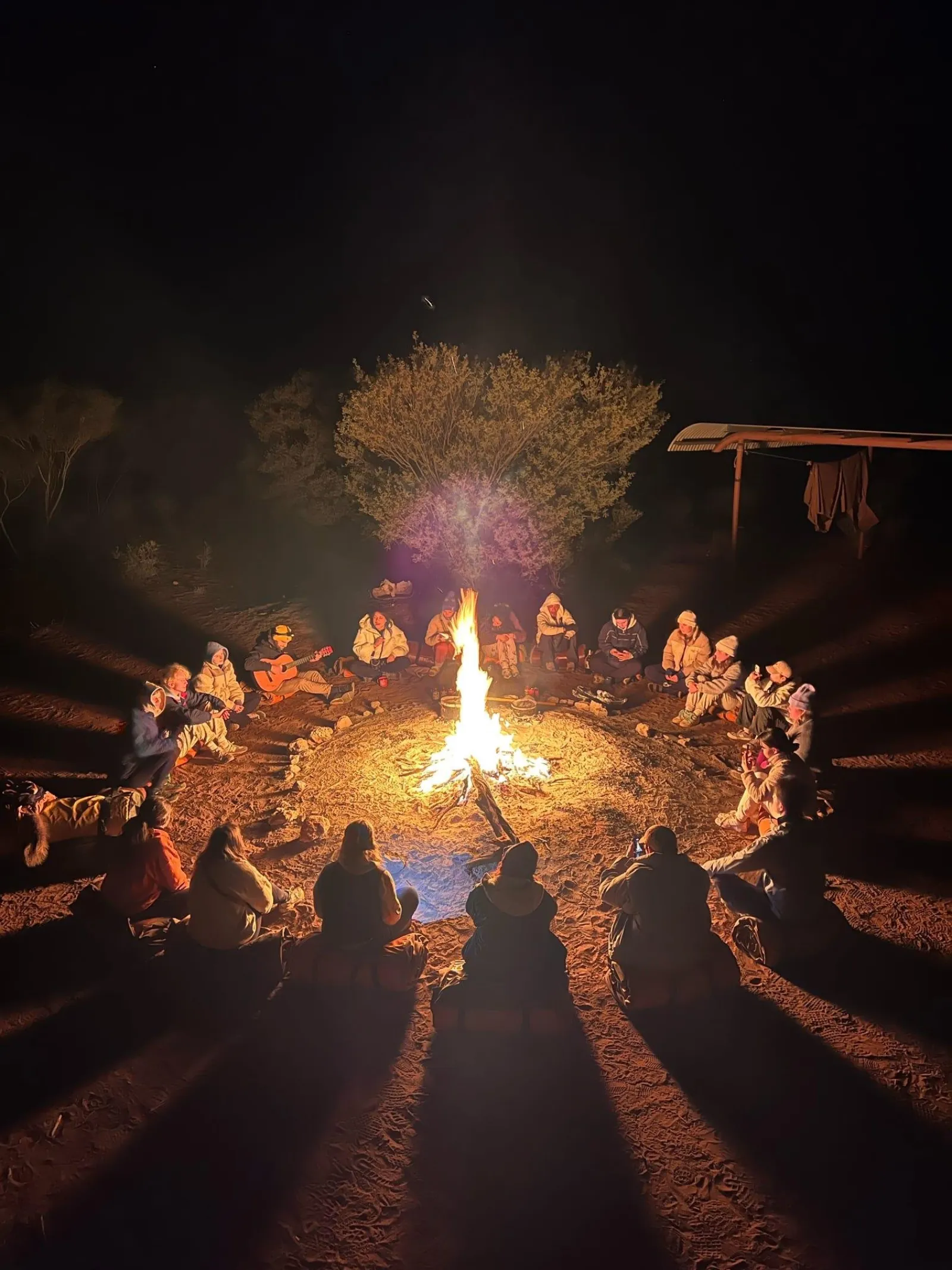 Travelers in warm jackets sitting in a circle around a blazing campfire on red sand at night